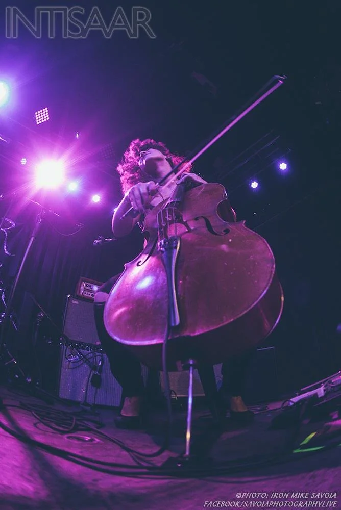 Natalie Mai Hall, a musician playing a cello on stage under purple and blue stage lighting, with audio equipment and cables in the background.