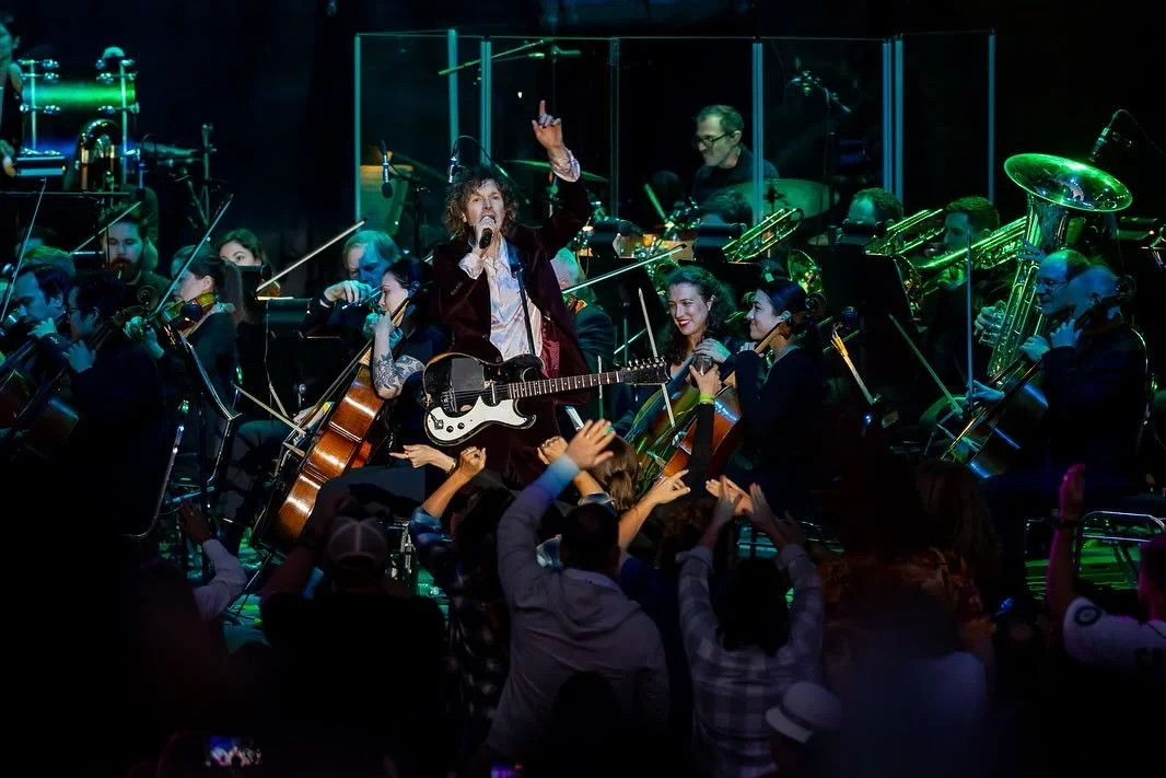 Musician with curly hair, guitar, and tattooed arm singing and performing in front of an orchestra, with audience applauding.