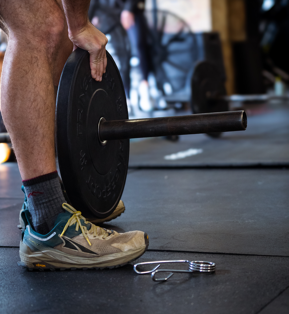 A person in athletic shoes and socks lifts a black weight plate with a barbell in a gym.