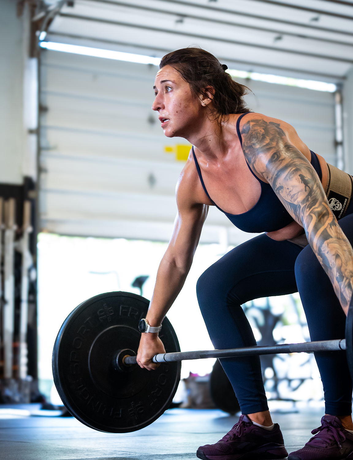 A woman with tattoos on her arm lifting a barbell in a gym.