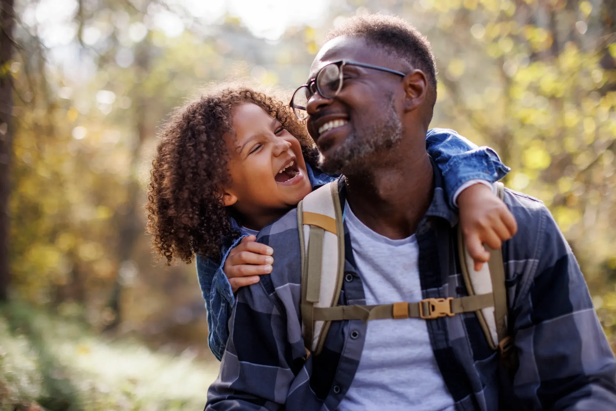A man with glasses and a beard giving a piggyback ride to a young girl with curly hair outdoors in a forested area during autumn, both smiling and laughing.