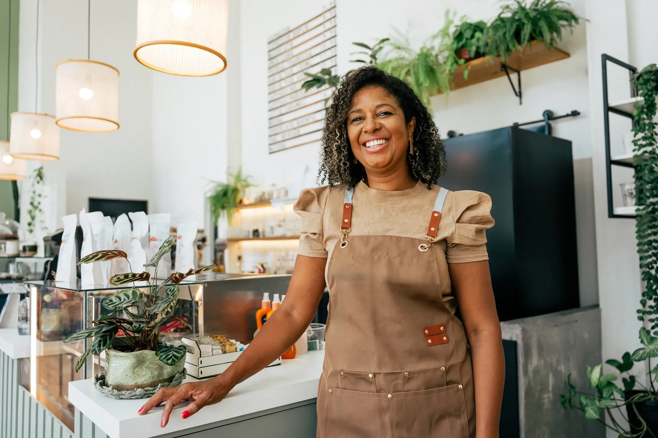 A woman standing behind a counter in a bright, modern shop, smiling at the camera. She is wearing a tan apron and a beige shirt with puffed sleeves. The shop has white walls, hanging pendant lights, and various plants.
