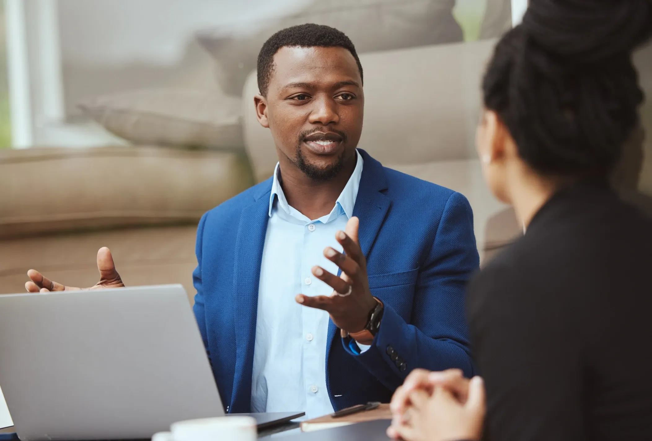 A man in a blue suit and light blue shirt speaking to a woman with dark hair in a black blazer during a business meeting.
