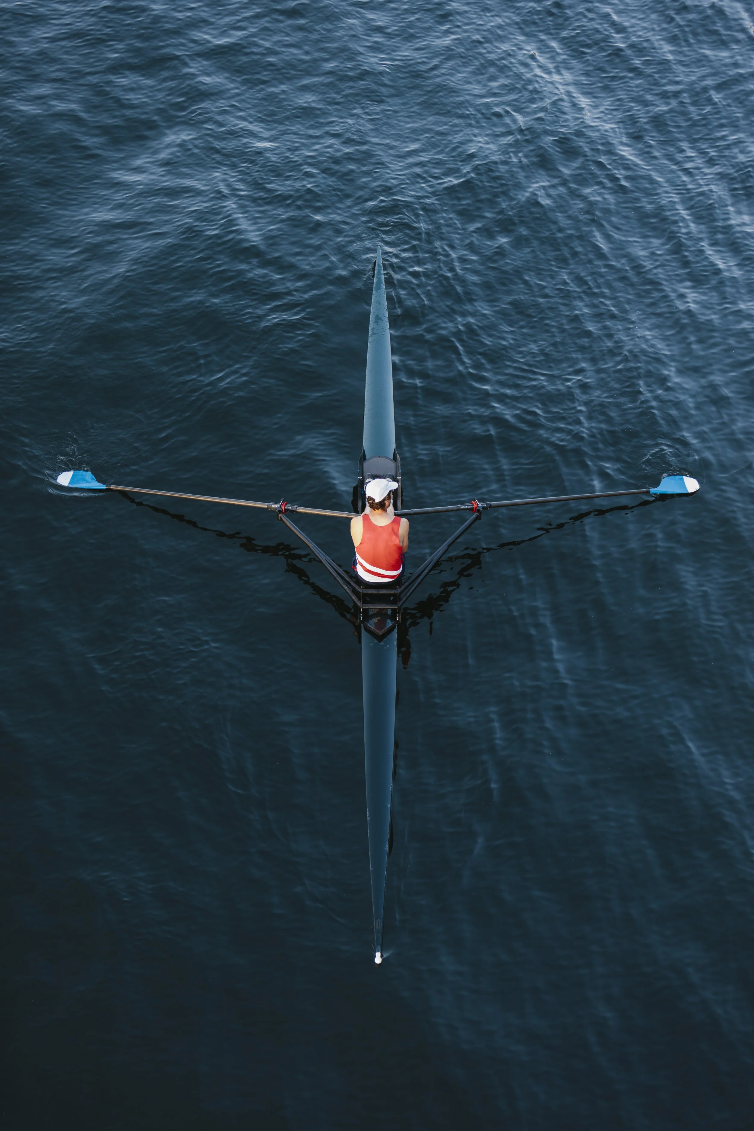 A rower, viewed from above, in a single scull wearing a red tank top and white hat is is surrounded by deep teal water
