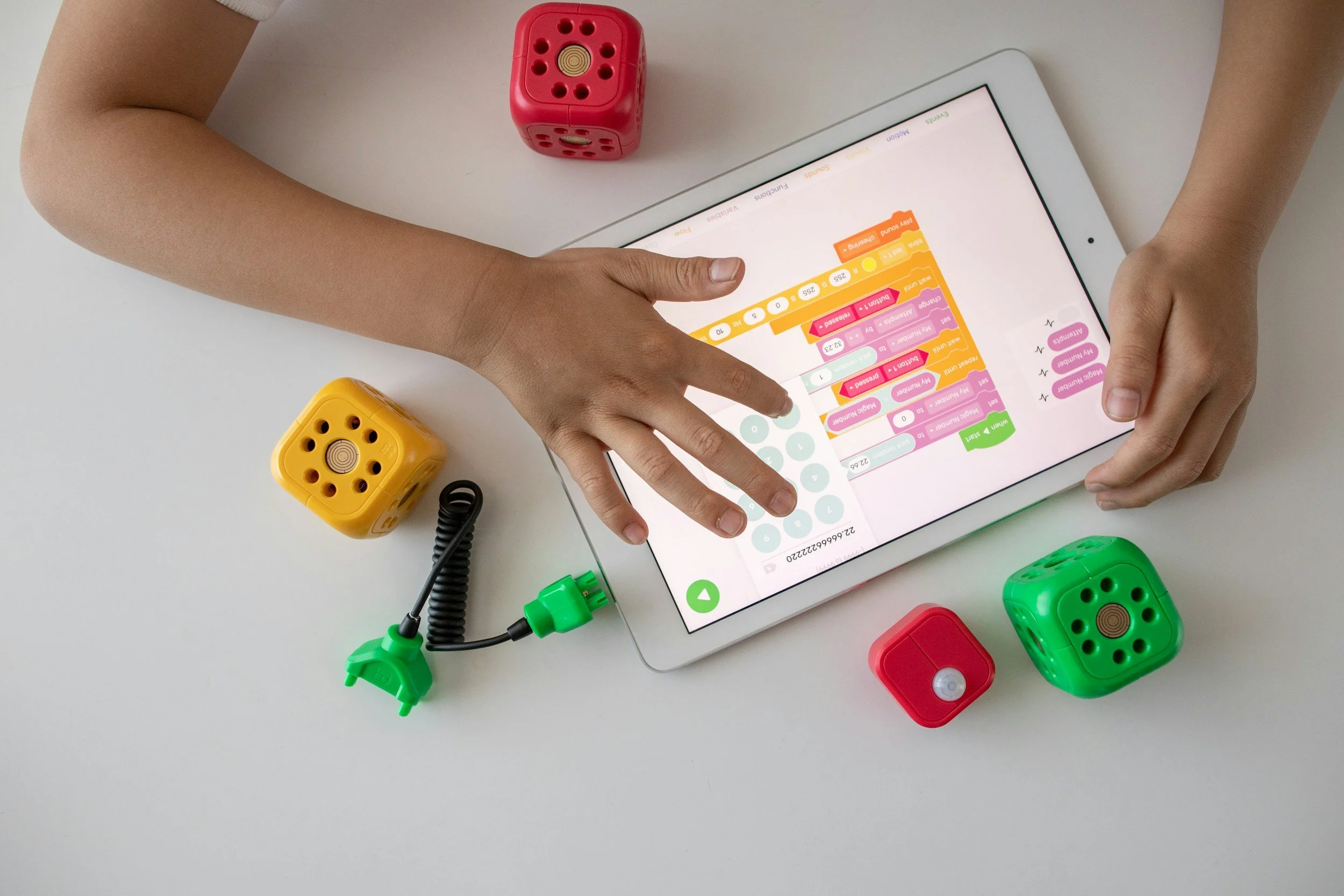 Child's hands interacting with a tablet displaying a visual programming interface, surrounded by colorful toy blocks and a cable.