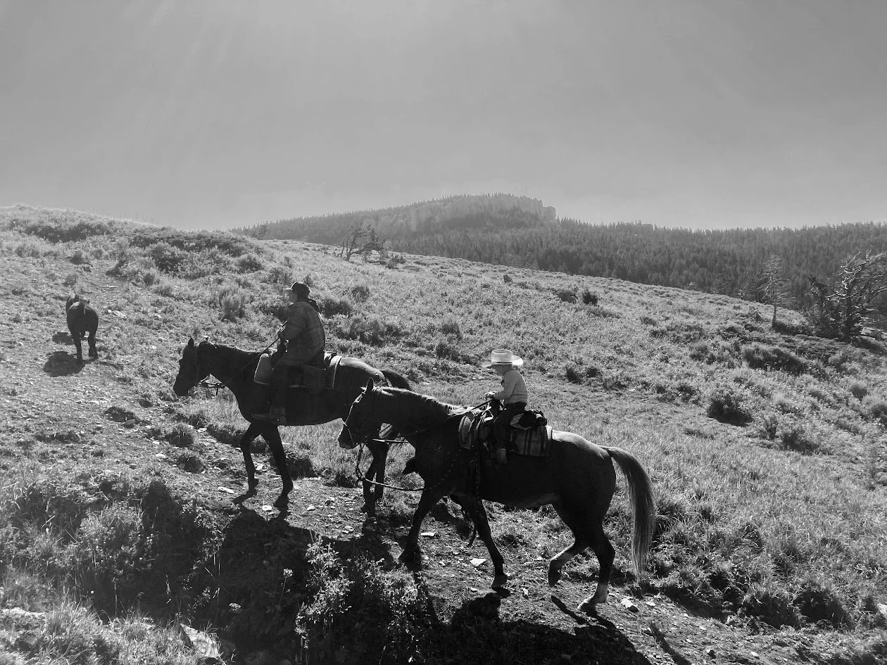 Two people riding horses on a trail in a natural landscape with hills and trees, black and white photo.