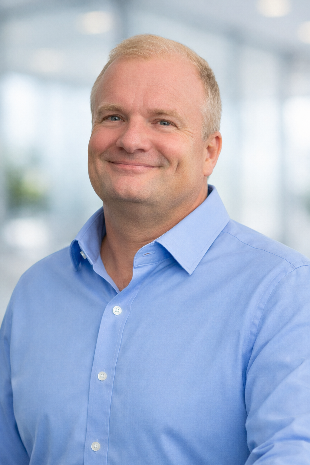 Portrait of a middle-aged man with short blond hair, blue eyes, smiling, wearing a light blue button-down shirt, in a bright, modern office environment.