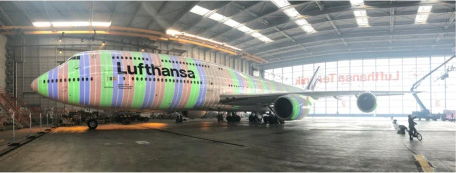 An indoor aircraft hangar with a Lufthansa airplane that has a colorful camouflage design with green, pink, purple, and blue stripes and the airline's logo on the fuselage.