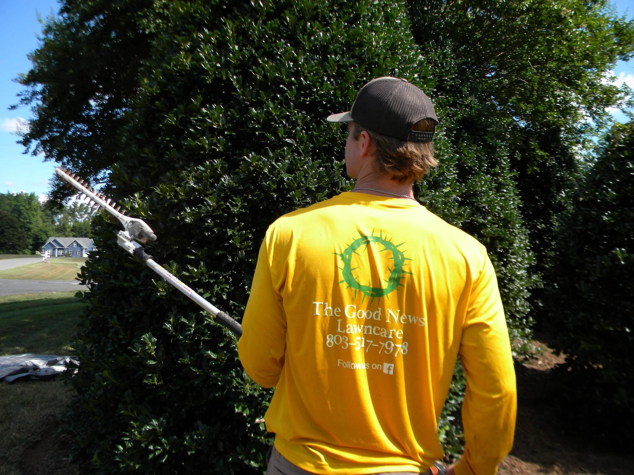 A man in a yellow shirt with the logo 'The Good News Lawns' and a phone number, holding landscaping trimmers, standing outdoors near a large green bush or tree.