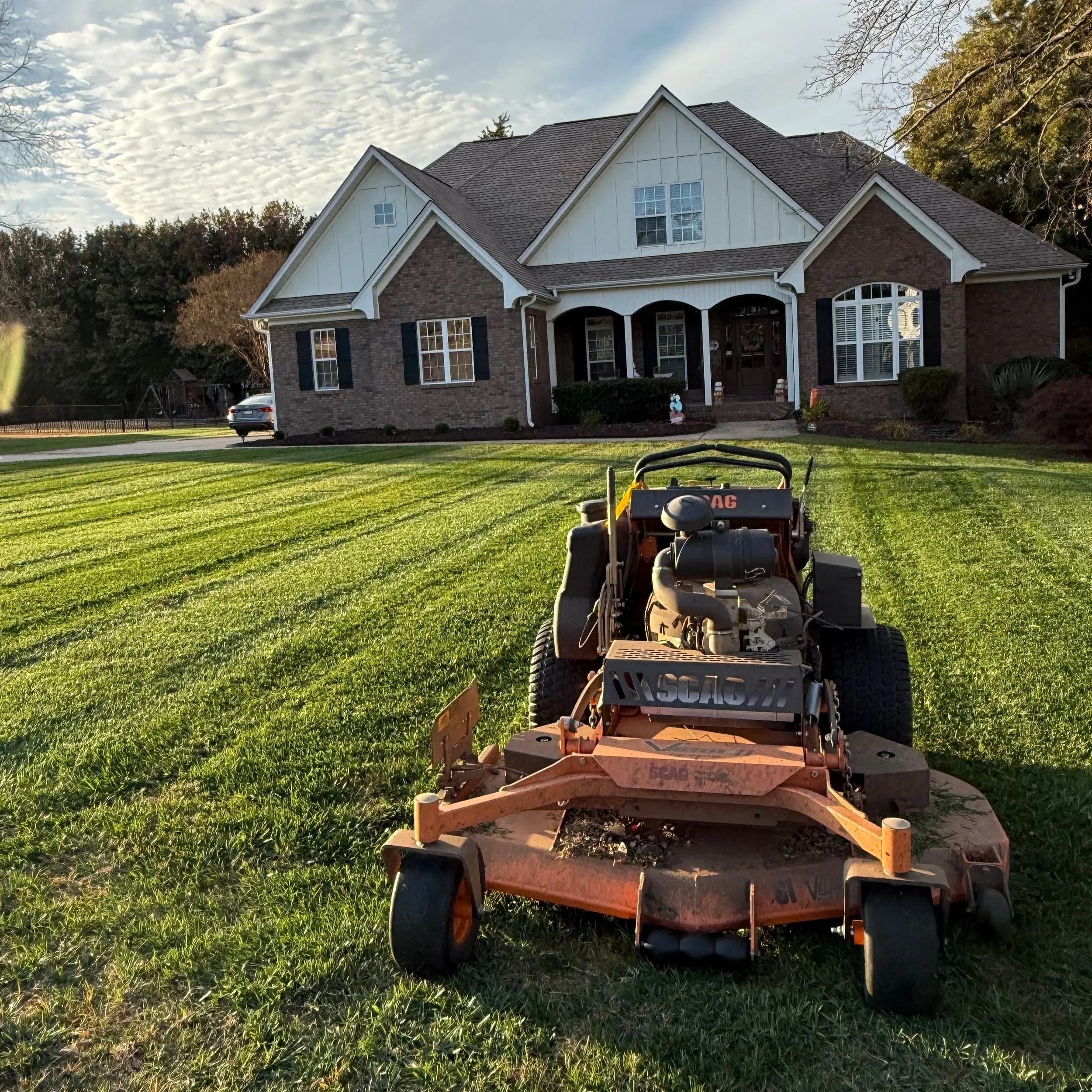 A lawn mower on freshly cut grass in front of a large brick house with white siding accents, windows, and a porch, under a partly cloudy sky.