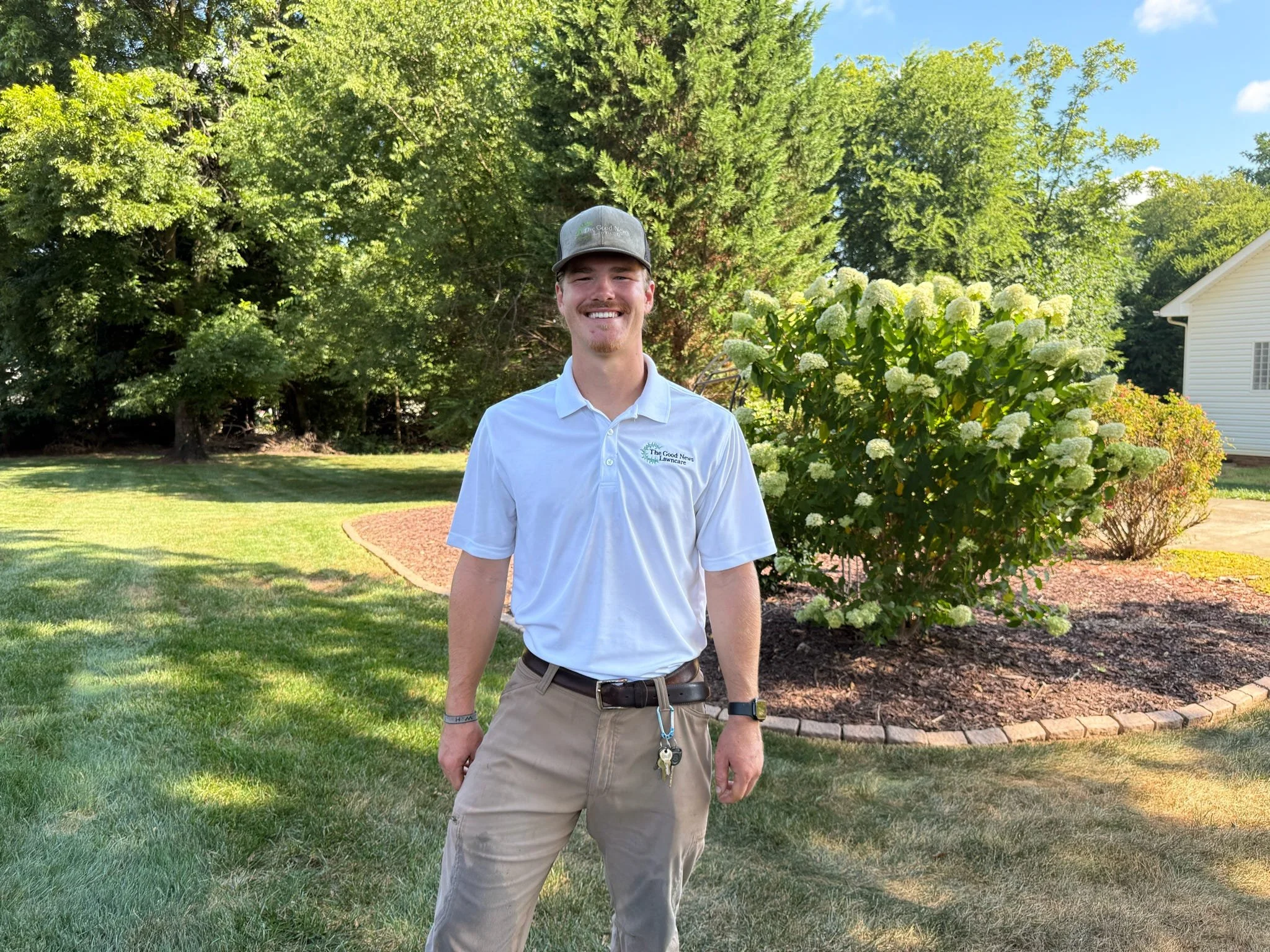 A smiling man in a white polo shirt and khaki pants standing on a lawn near a flower bush.