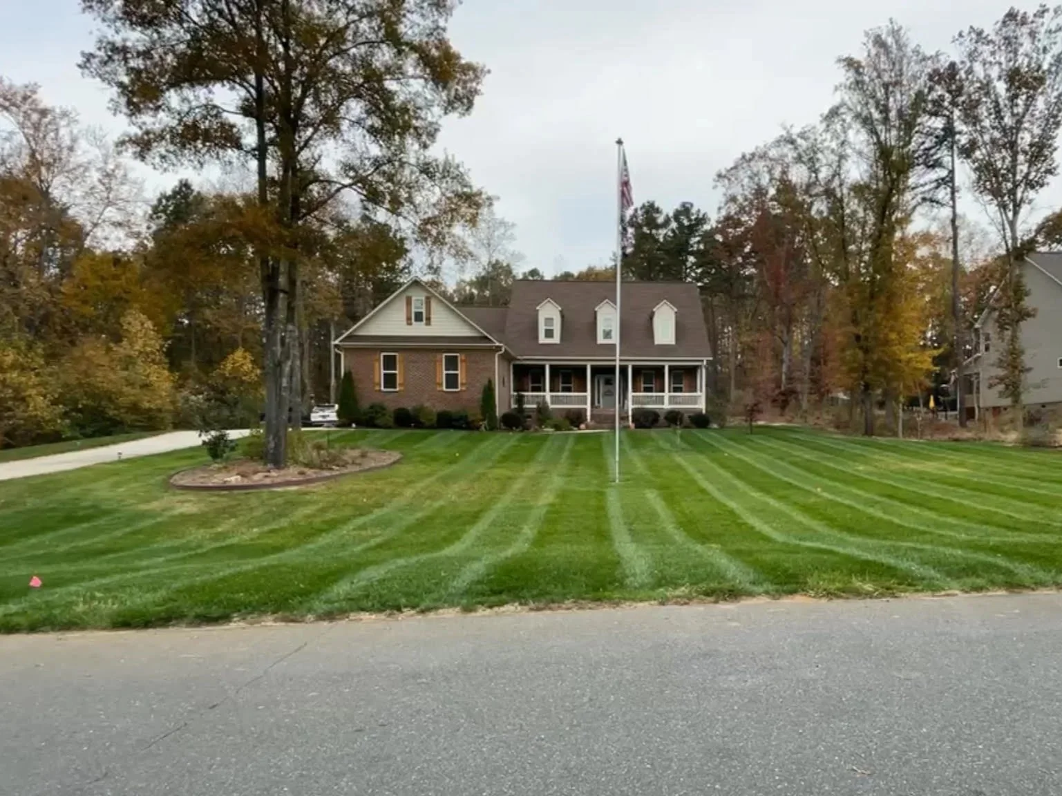 A house with a well-maintained lawn, a flagpole with an American flag, and a tree in front. The house has brick and siding exterior, surrounded by trees with autumn-colored leaves.