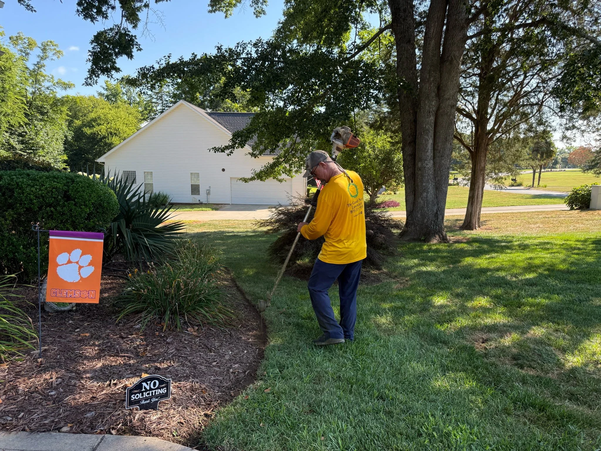 A man in a yellow shirt and cap is gardening in a residential yard, using a rake near a flower bed with a Clemson University sign. There are trees, manicured grass, and a white house in the background.