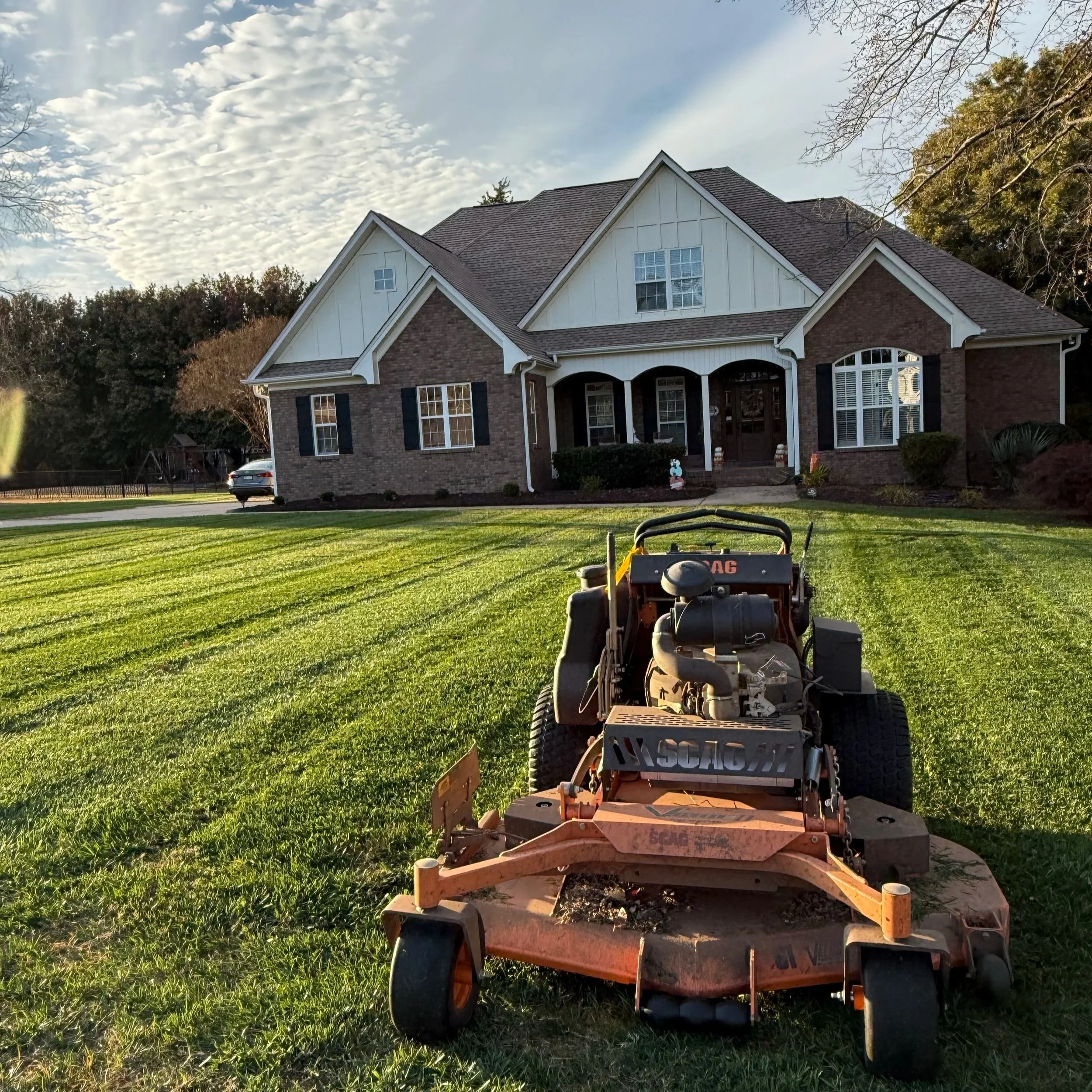 A residential house with a well-maintained lawn, a large mower in the foreground, and trees in the background, under a partly cloudy sky.