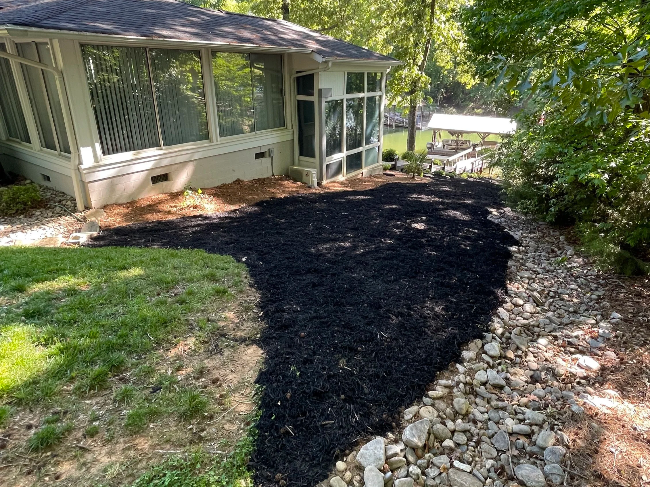 Backyard with new black mulch border around garden beds, a house with large windows, and a lakeside dock with a shed in the background.