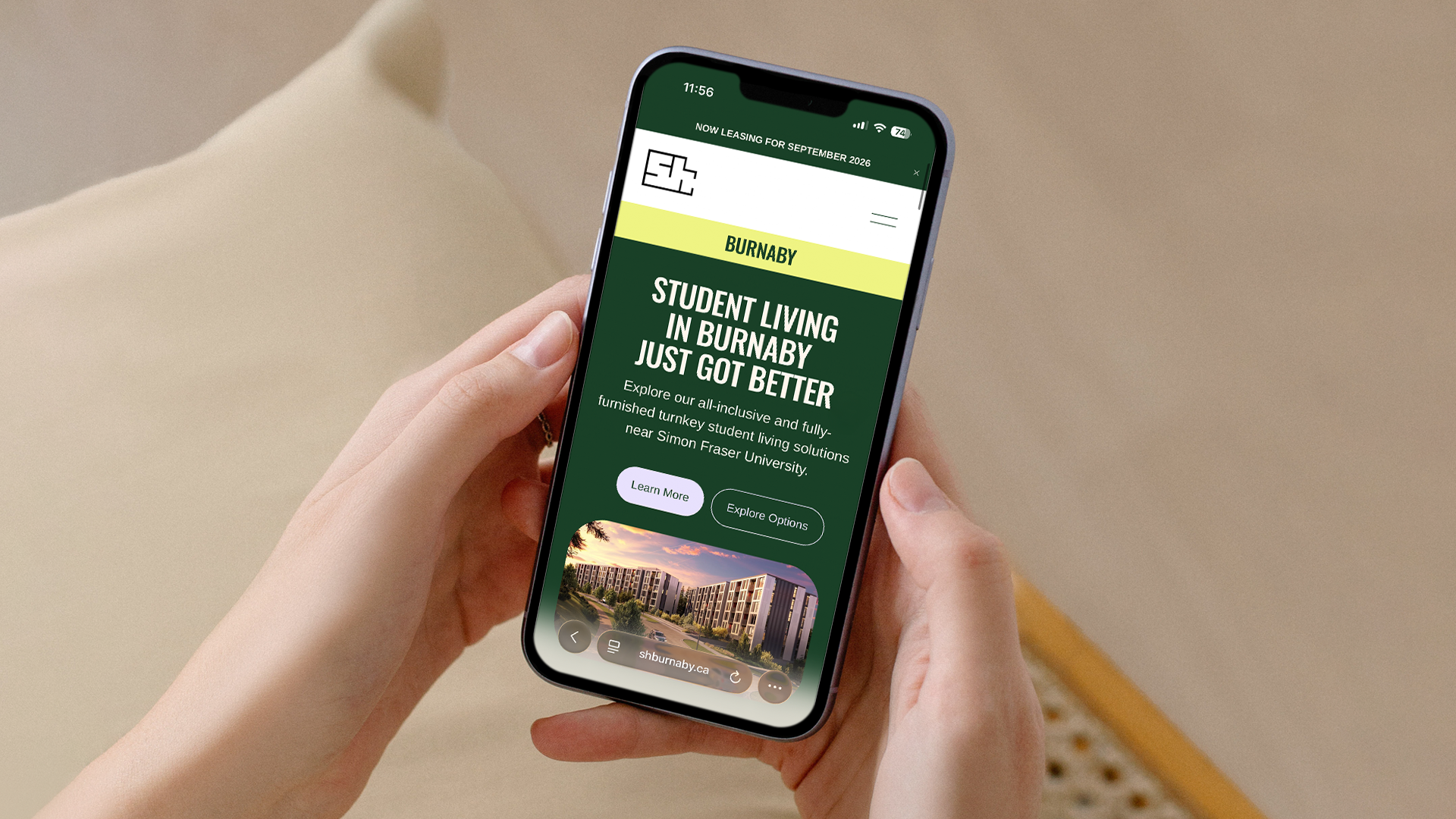 Close-up of a person holding a smartphone displaying a website about student living in Burnaby, with a building picture at the bottom and buttons labeled 'Learn More' and 'Explore Options'.