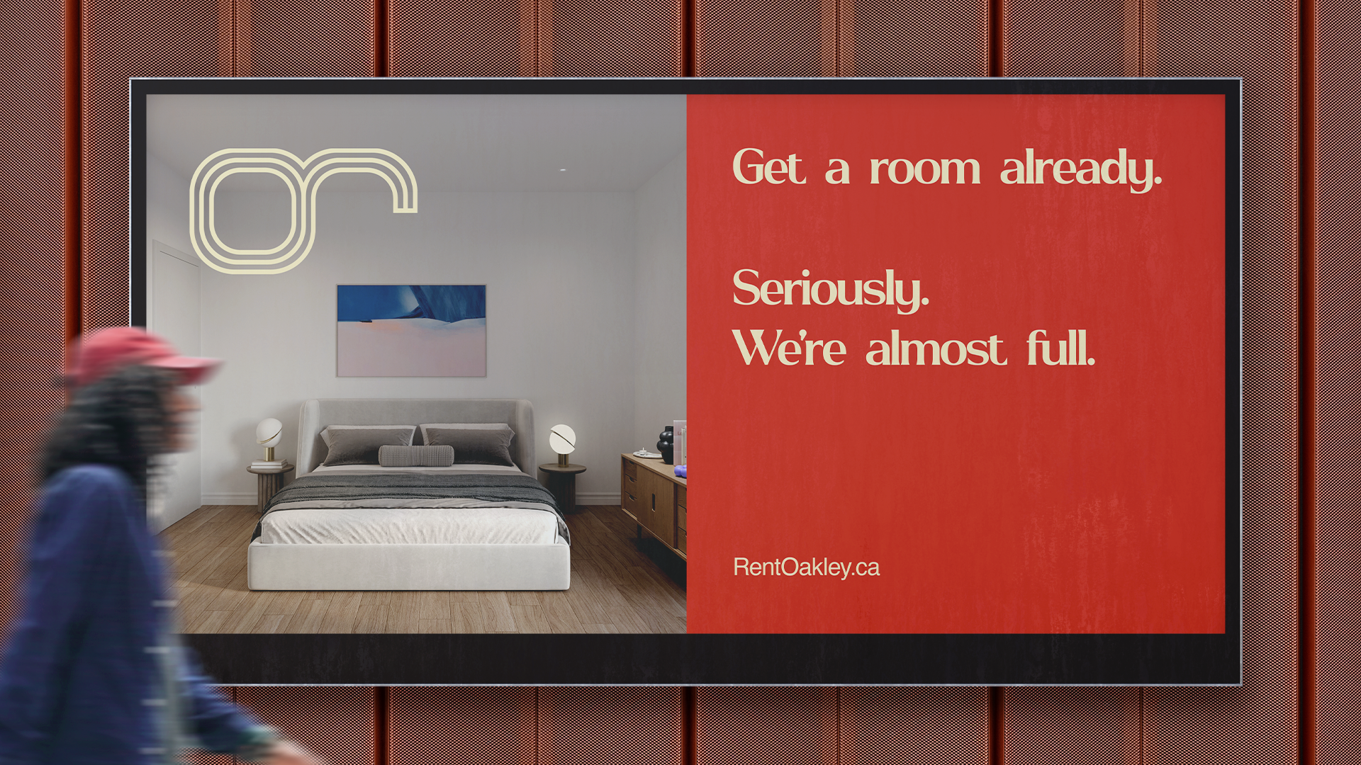 Billboard advertisement for RentOakley.ca featuring an interior view of a modern bedroom with a bed, bedside tables, lamps, and a painting on the wall, with text that reads 'Get a room already. Seriously. We're almost full.'