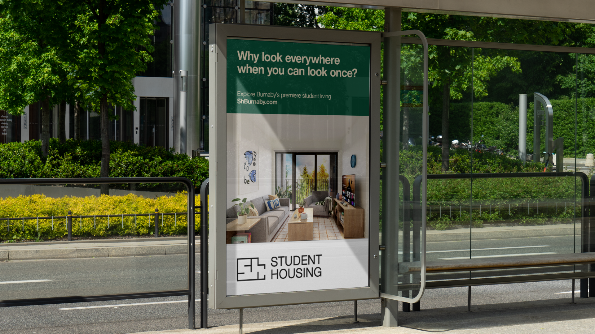 Bus stop advertisement for student housing featuring a living room interior with couches, a coffee table, and a TV, set outside with green trees and a street in the background.