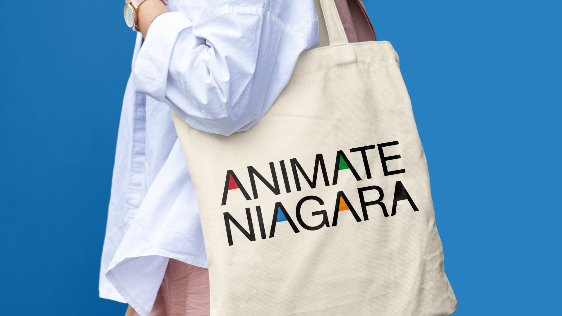 A person wearing a white shirt carrying a canvas tote bag with the logo 'ANIMATE NIAGARA' in colorful letters, standing against a solid blue background.