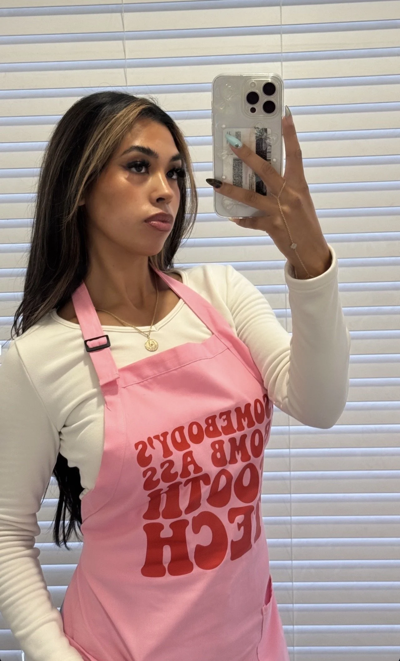Woman taking a mirror selfie with a smartphone, wearing a white long-sleeve shirt and a pink apron with red text, in front of closed blinds.