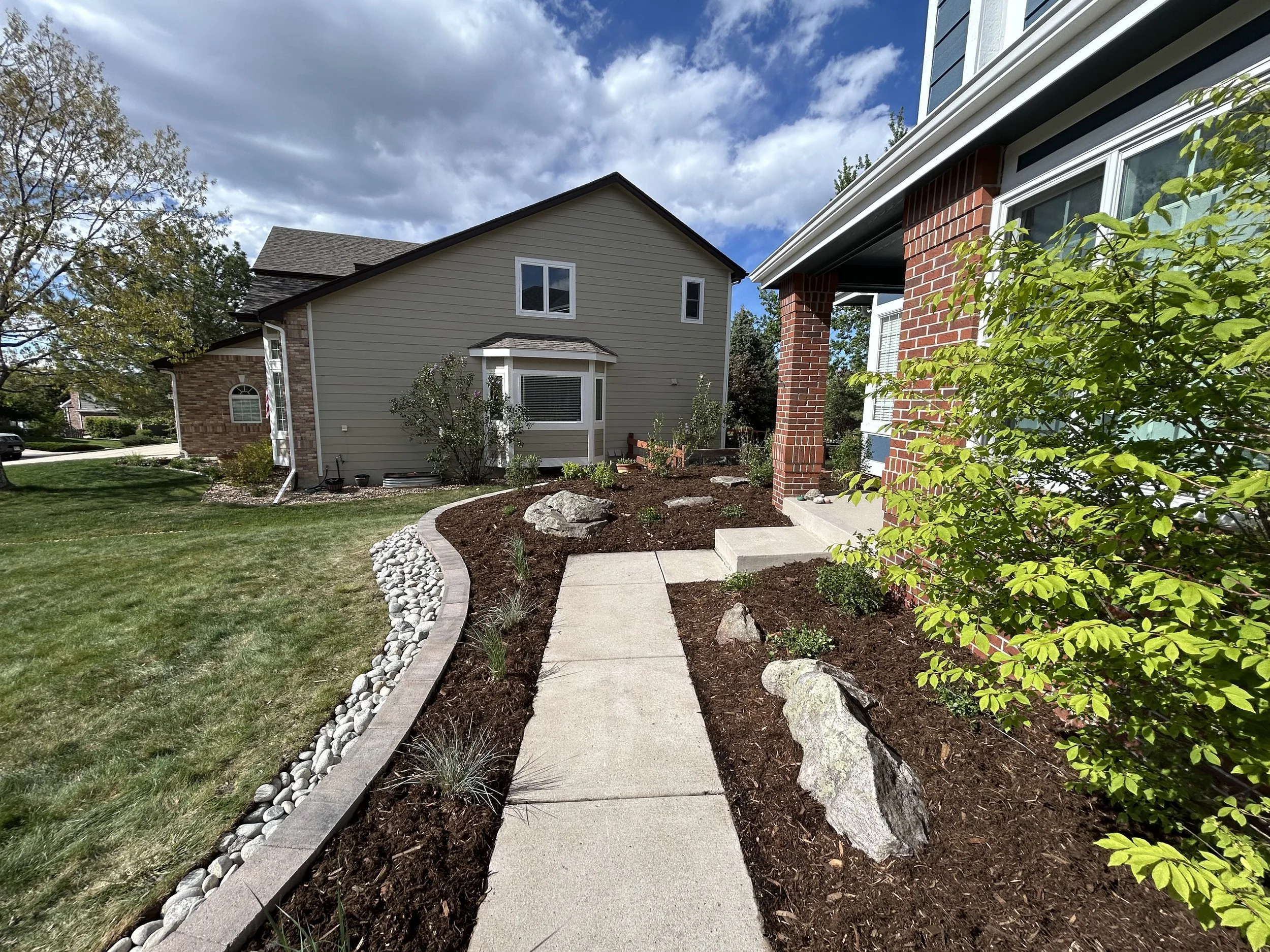 A paved sidewalk leading to a porch of a house, with landscaped area with rocks, soil, and small plants on either side. The house has brick and siding exterior, and other houses are visible in the background under a partly cloudy sky.
