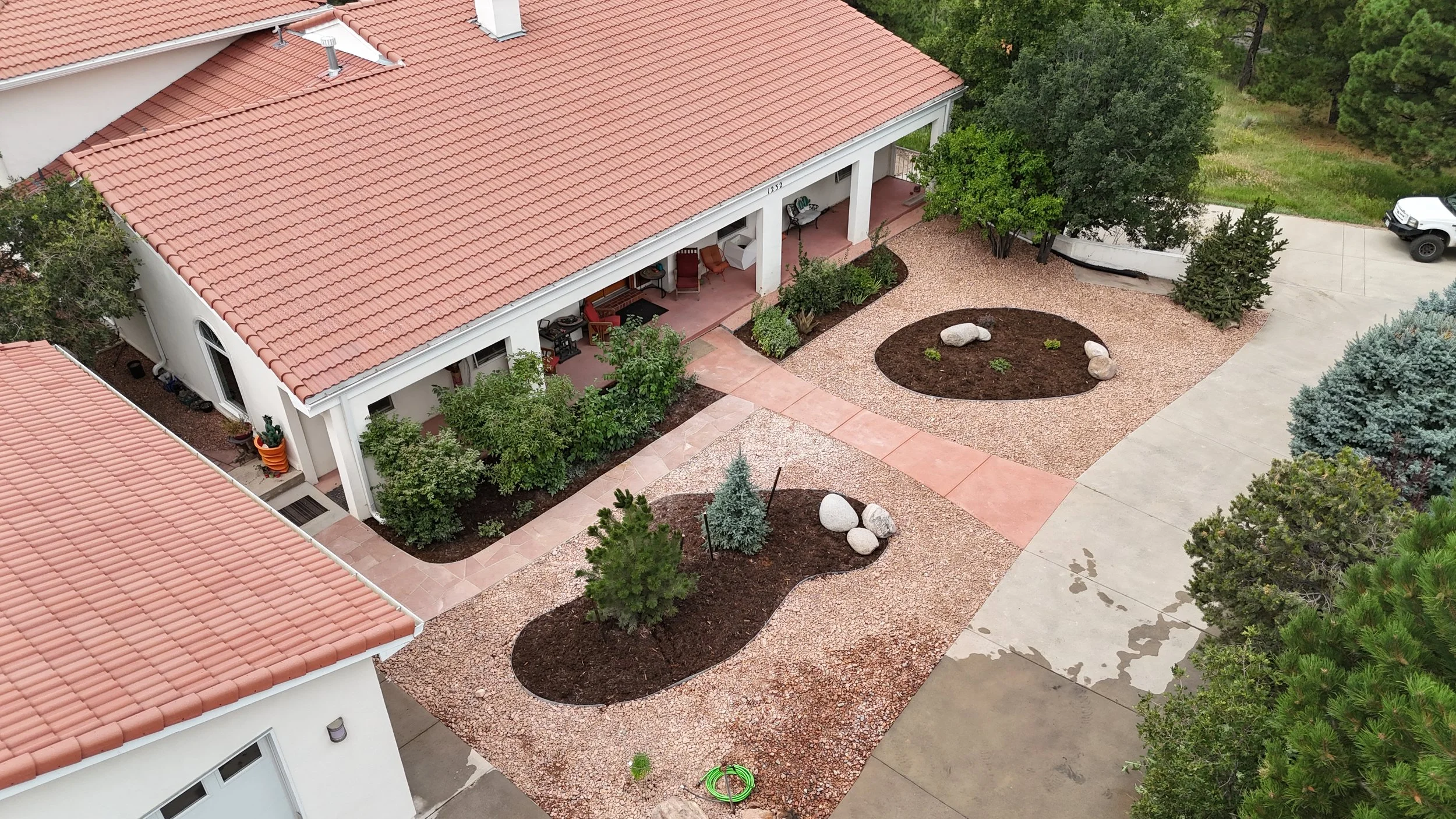 An aerial view of a residential backyard with a house featuring a red-tiled roof, a covered porch with outdoor furniture, and landscaped areas with trees, bushes, rocks, and a concrete driveway.