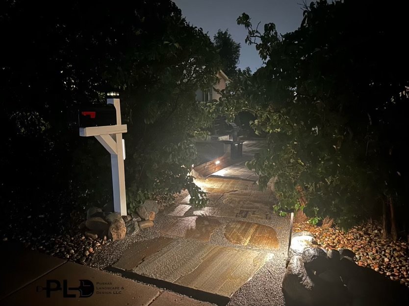 A walkway at night with rain, illuminated by outdoor lights, tree branches hanging over the path, a black mailbox on a post to the left, and rocks lining the edges.