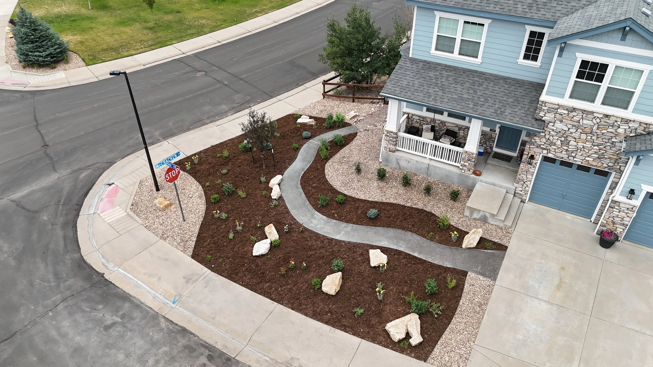 An aerial view of a corner of a residential area showing a light blue house with a stone accent wall, a small porch with chairs, a landscaped front yard with a curved gravel pathway, young bushes, and rocks, and a street with a stop sign at the corner.