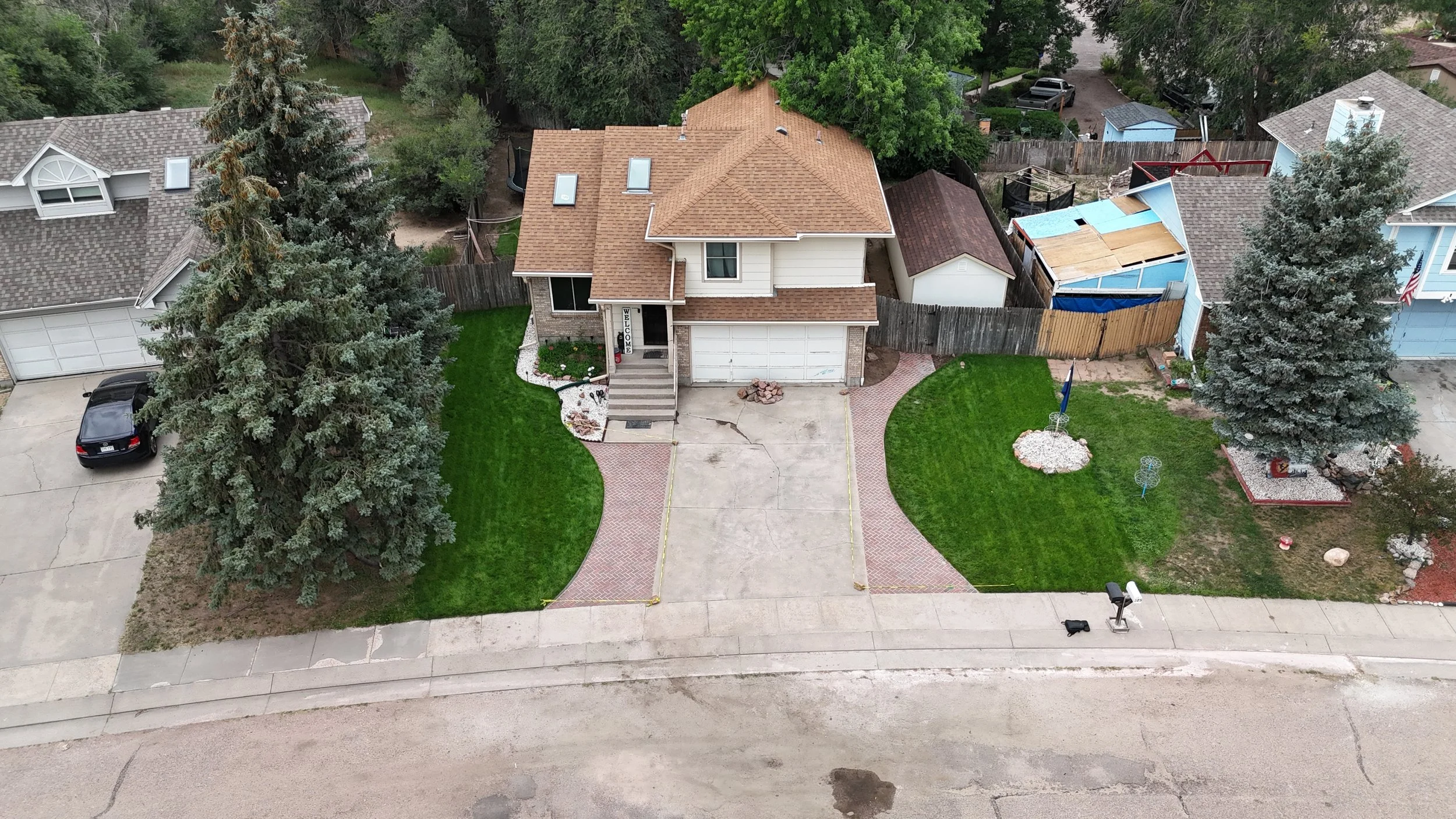 Aerial view of a suburban house with a two-car garage, a front porch with stairs, landscaped yard with grass, trees, and decorative rocks, neighboring houses, and a sidewalk.