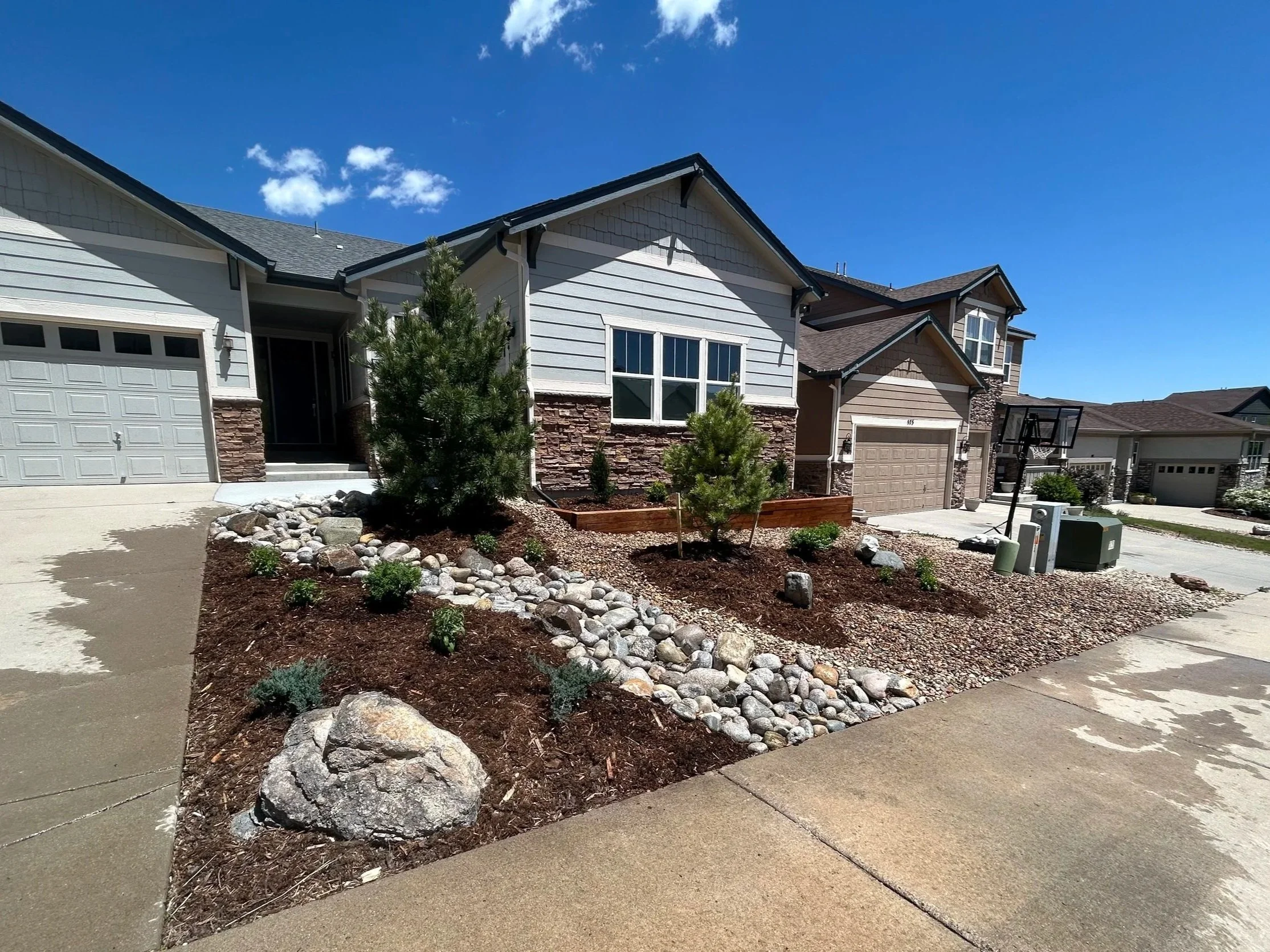 Front view of a suburban house with a landscaped yard, small trees, rocks, and mulch, under a clear blue sky.