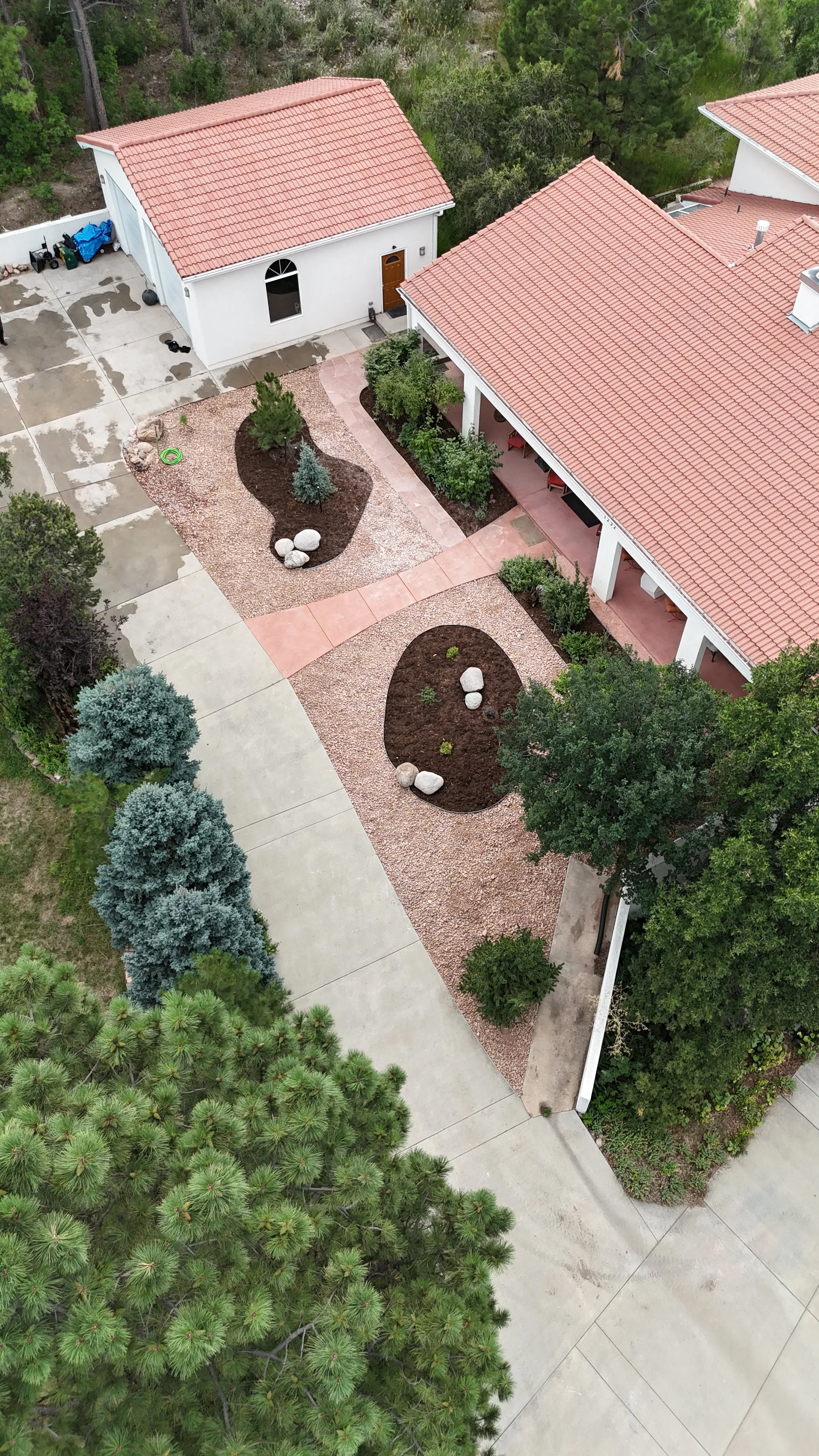 An aerial view of a residential backyard with two white houses with red tile roofs, landscaped with gravel, bushes, trees, and rocks, and a concrete driveway.