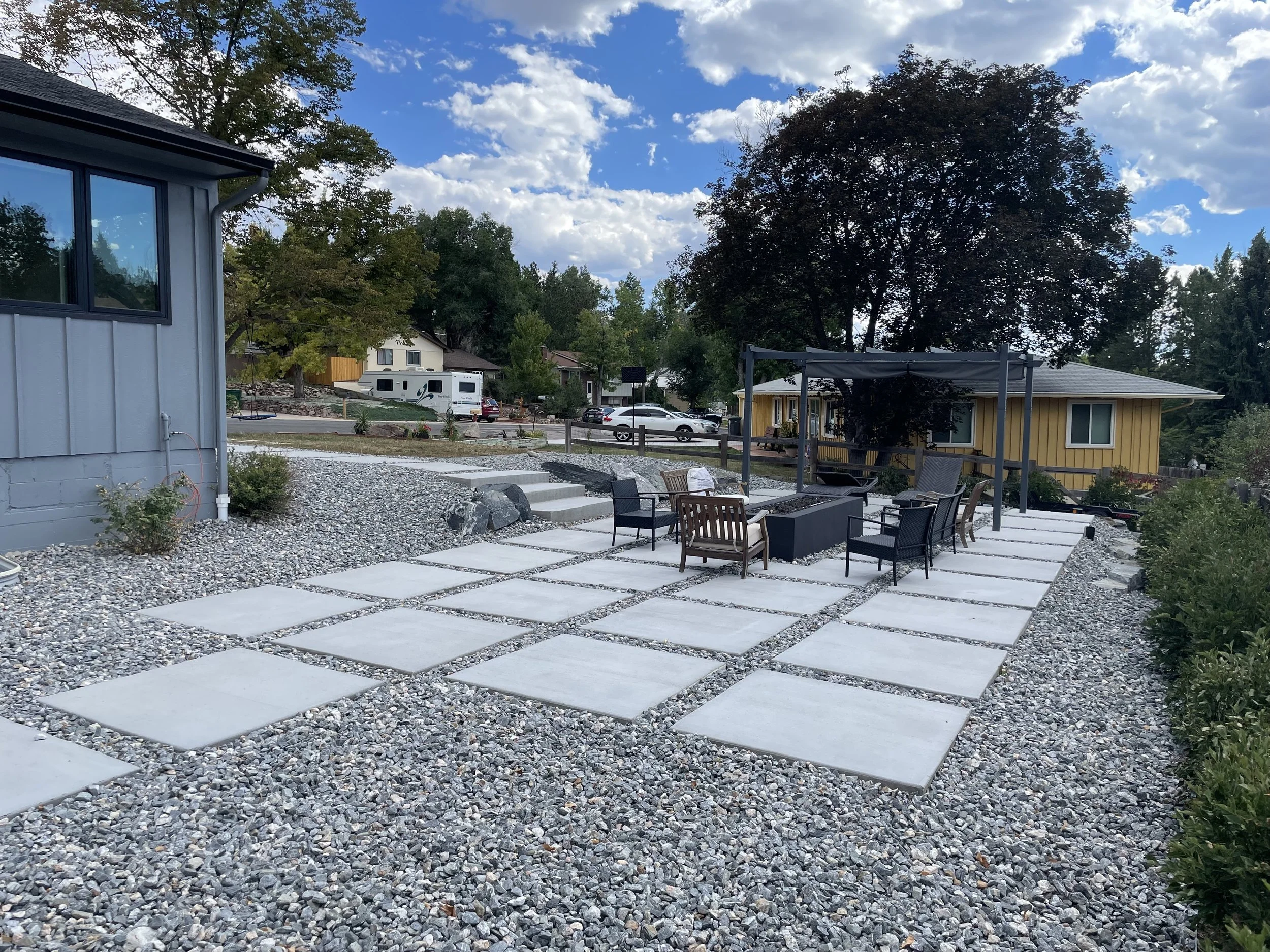 Backyard patio with large concrete pavers, gravel ground cover, outdoor seating including chairs and a firepit, a shaded structure, surrounded by trees and houses, under a partly cloudy sky.