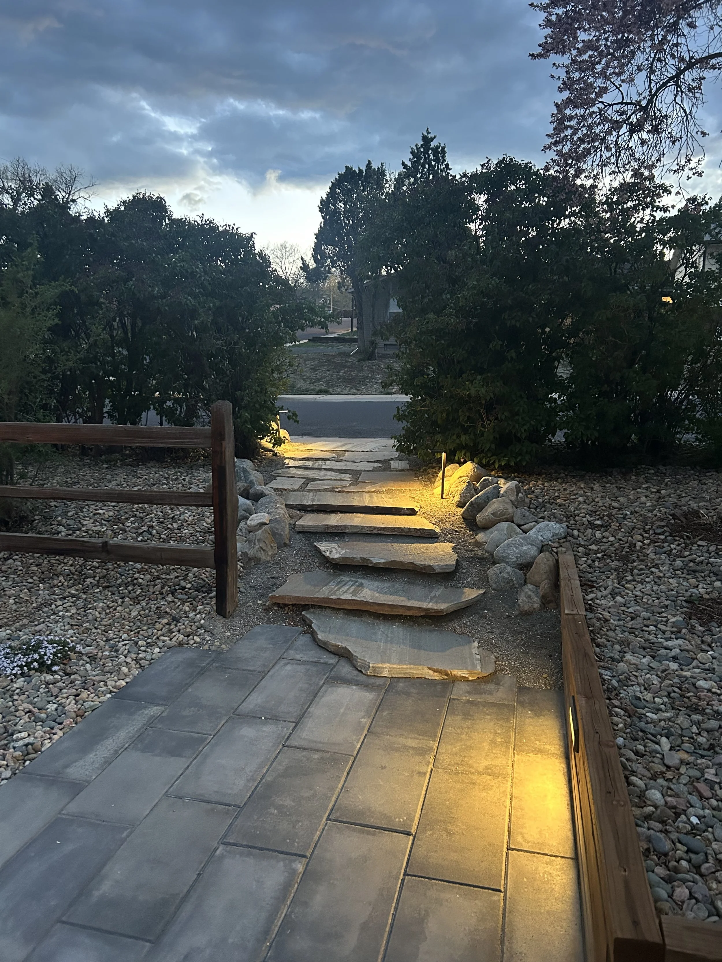 A pathway with large, flat stepping stones and bordered by rocks, illuminated by small ground lights in front of bushes and trees during twilight.