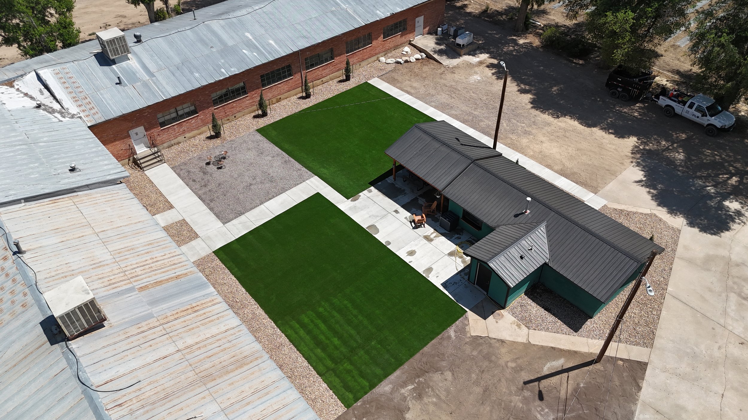 An aerial view of a building with a metal roof and a small yard, featuring two sections of artificial green grass, a gravel area with a few chairs, a small covered patio with outdoor furniture, and surrounding trees and parked vehicles.