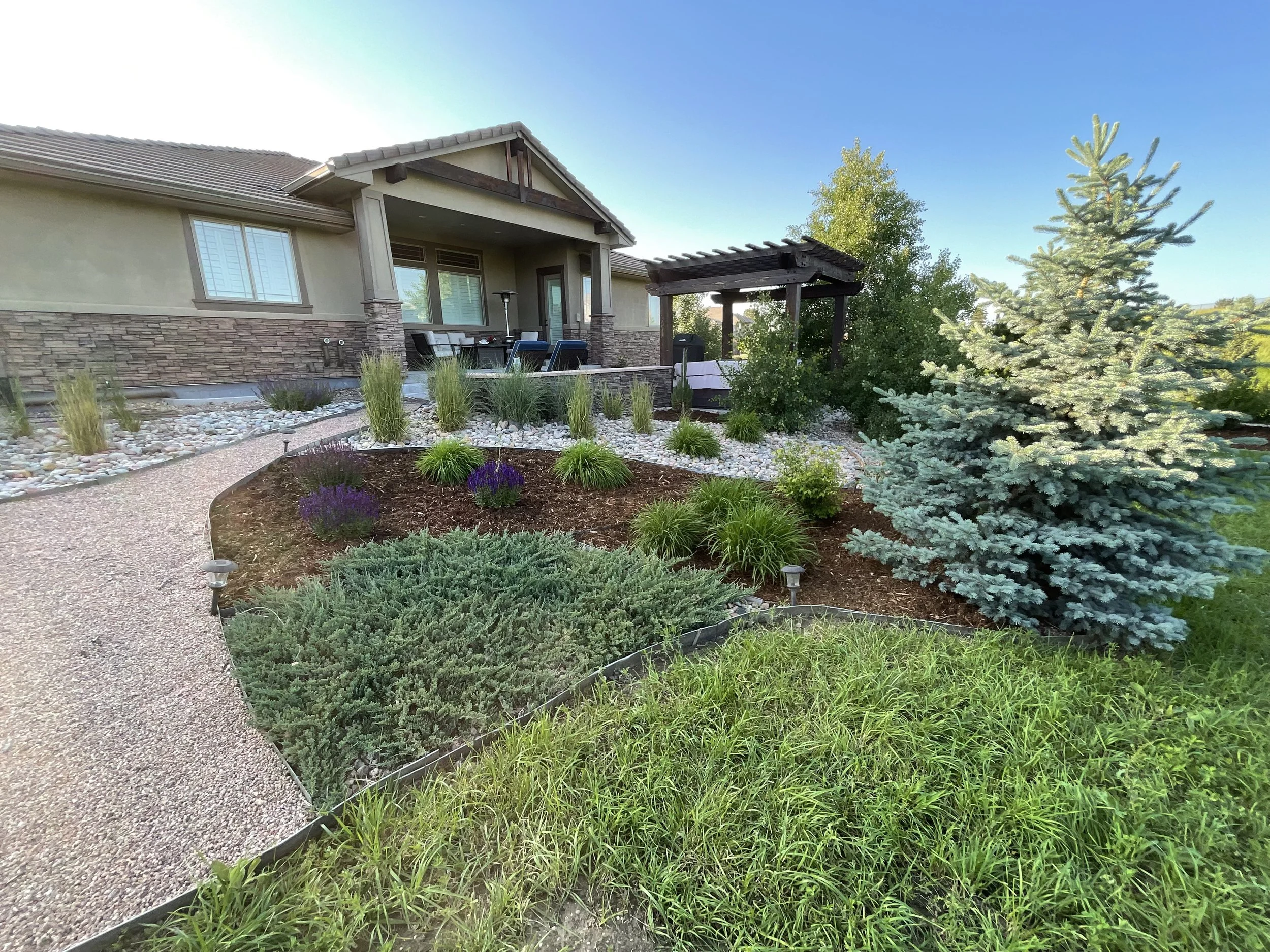 A landscaped backyard with a walking path, green plants, trees, shrubs, a patio with outdoor furniture, and a house with stone and stucco exterior during daytime.
