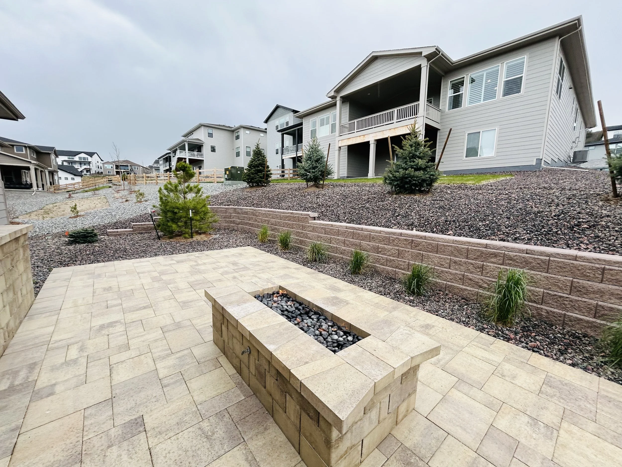 Backyard with beige tiled patio, decorative fire pit, small trees, and a tiered landscape with gravel and brick retaining wall in a suburban neighborhood.