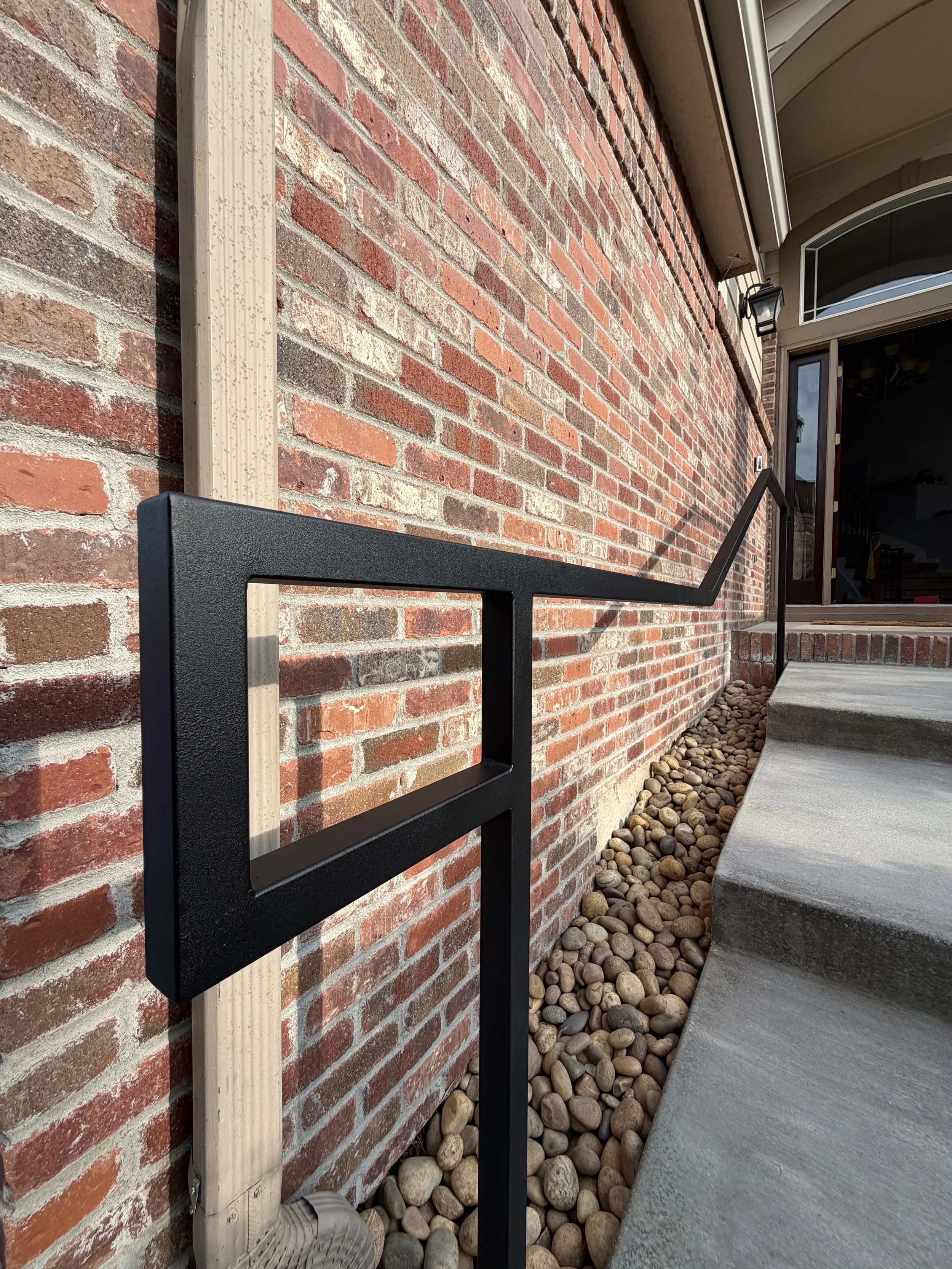 Close-up of a brick house exterior with a stucco column, a black metal handrail, a concrete step, rocks along the foundation, a black outdoor wall lantern, and an open doorway leading inside.