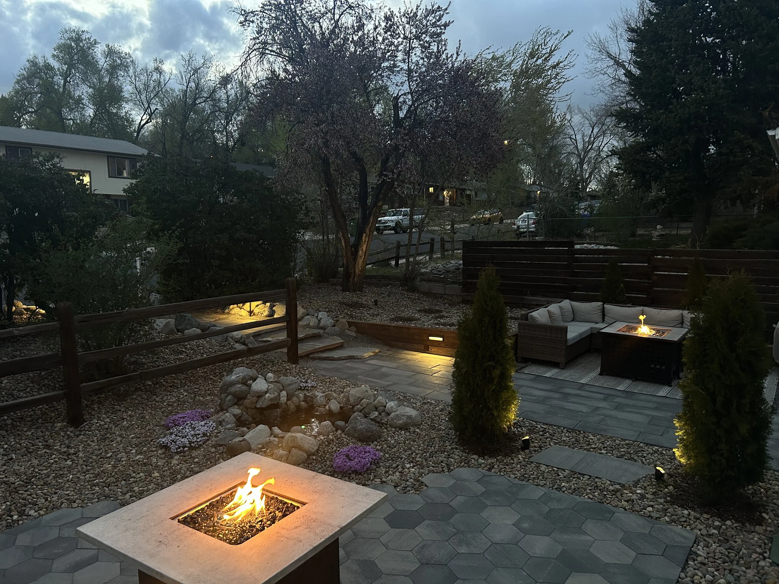 An outdoor patio area with fire pits, a stone wall, small shrubs, and a courtyard with a wooden fence and trees at dusk.