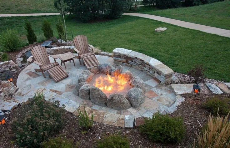 Backyard fire pit area with two wooden lounge chairs and a small table, surrounded by rocks and decorative stone wall, with a well-maintained grassy lawn and walkway in the background.