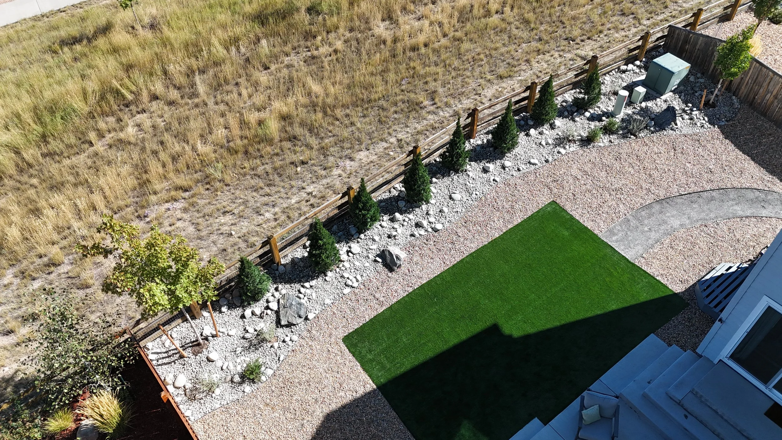 An aerial view of a backyard showing a green grass patch, concrete pathway, gravel area with small bushes and trees, a garden bed with rocks, and a wooden fence separating the yard from a dry grassy field.