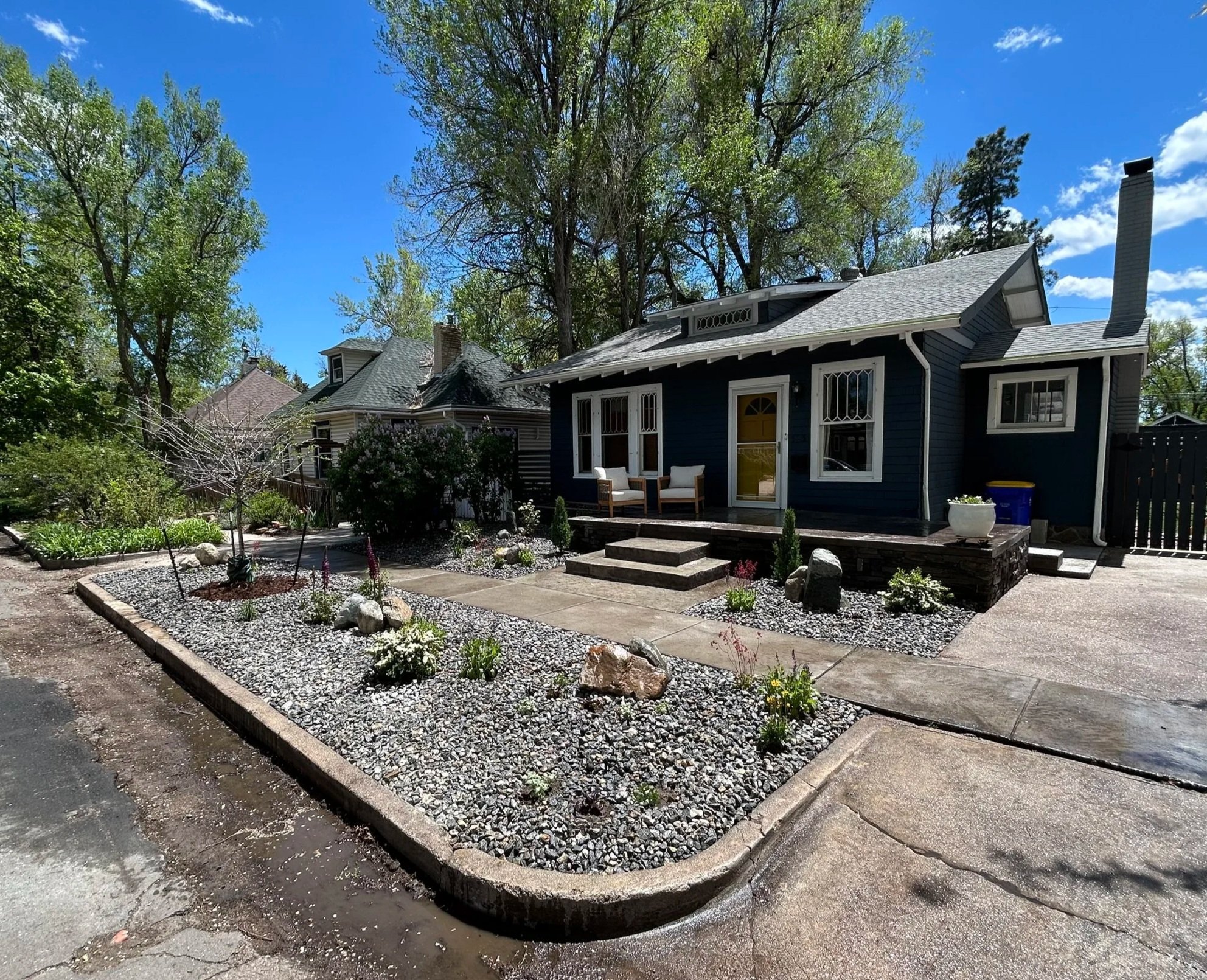 A well-maintained house with a small front porch, surrounded by a landscaped yard with rocks and various small plants, trees, and shrubs, under a blue sky with scattered clouds.