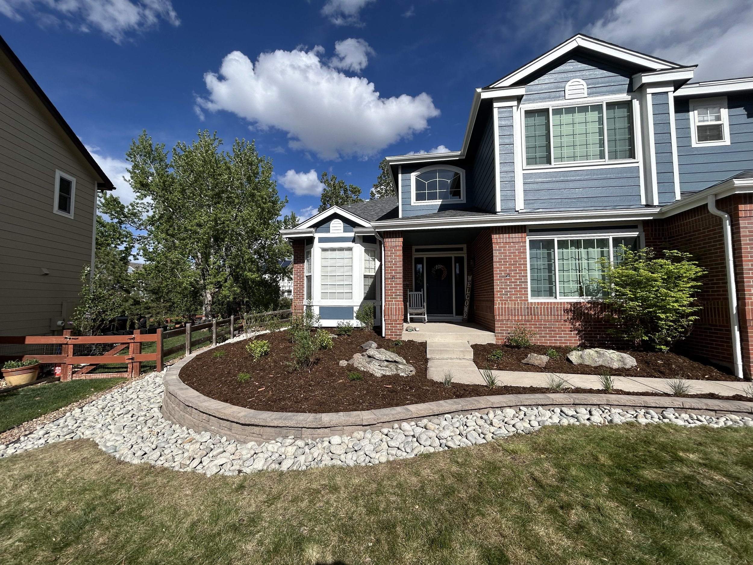 Front yard of a blue and brick house with landscaped garden, pathway, and trees under a partly cloudy sky.