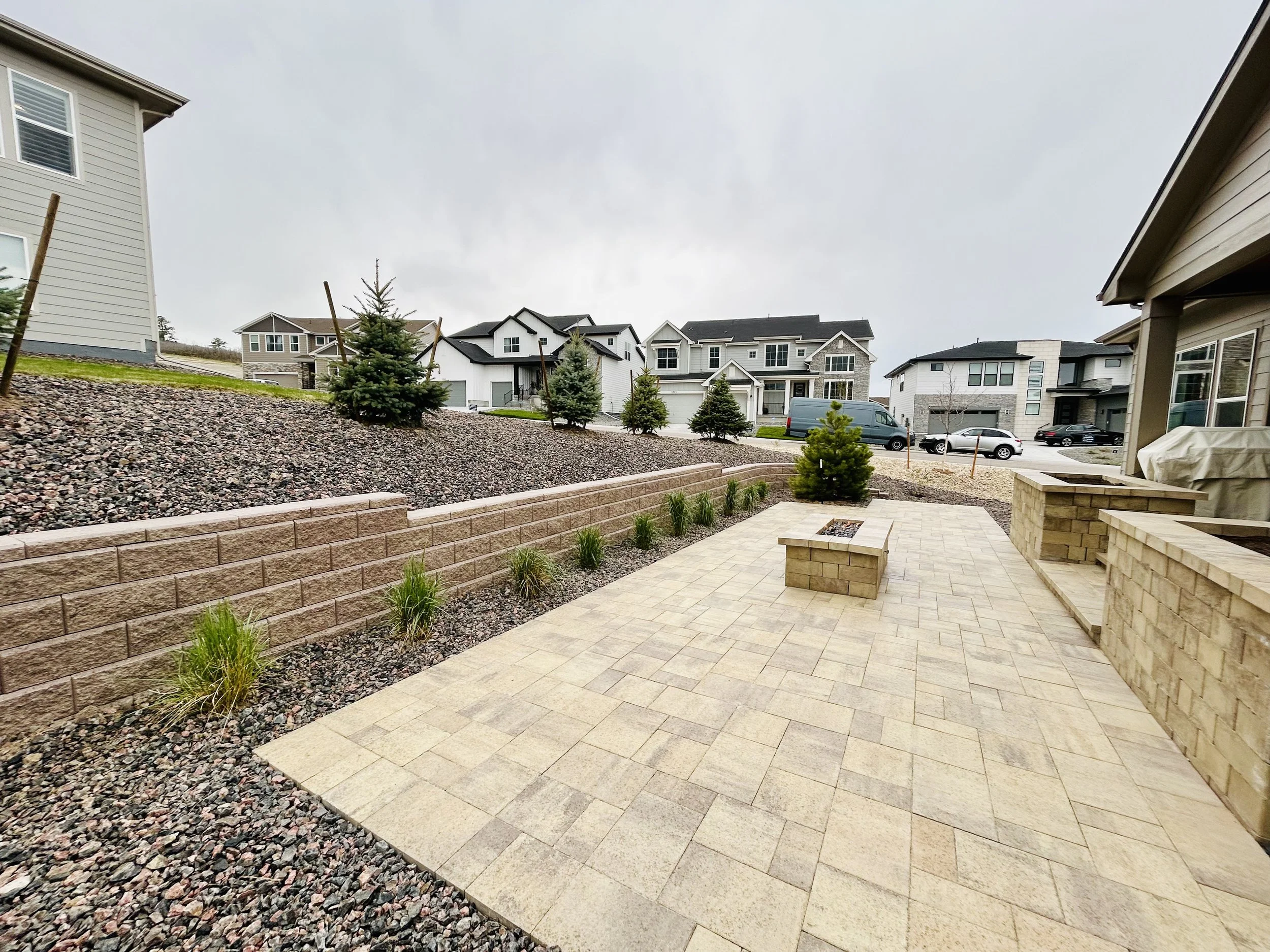 Backyard patio with beige paver stones, low brick walls, small conifer trees, and a fire pit, in a suburban neighborhood with modern houses and a cloudy sky.