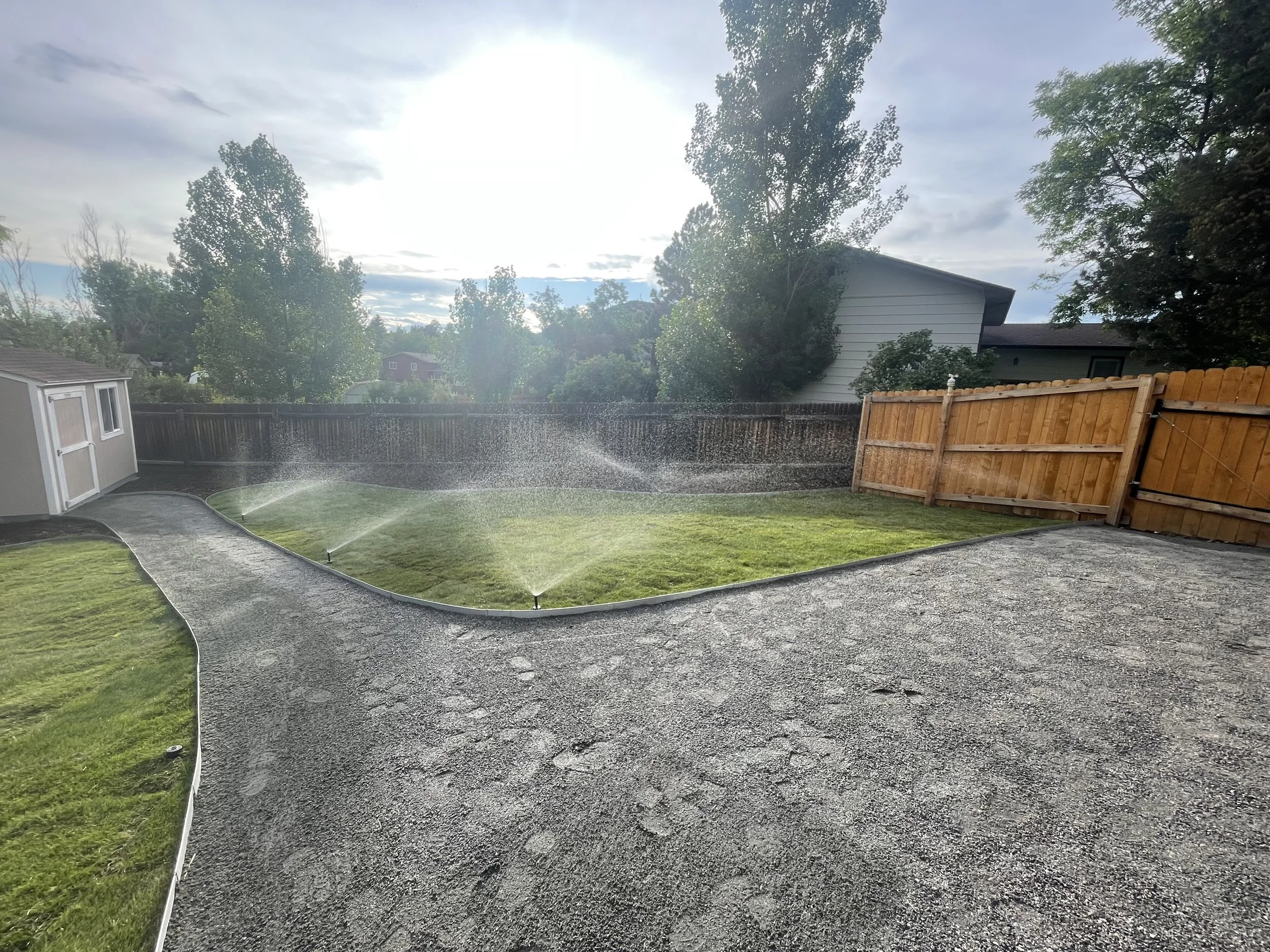 A backyard with a gravel patio, a lawn being watered by sprinklers, trees, and a wooden fence with a small shed on the left.