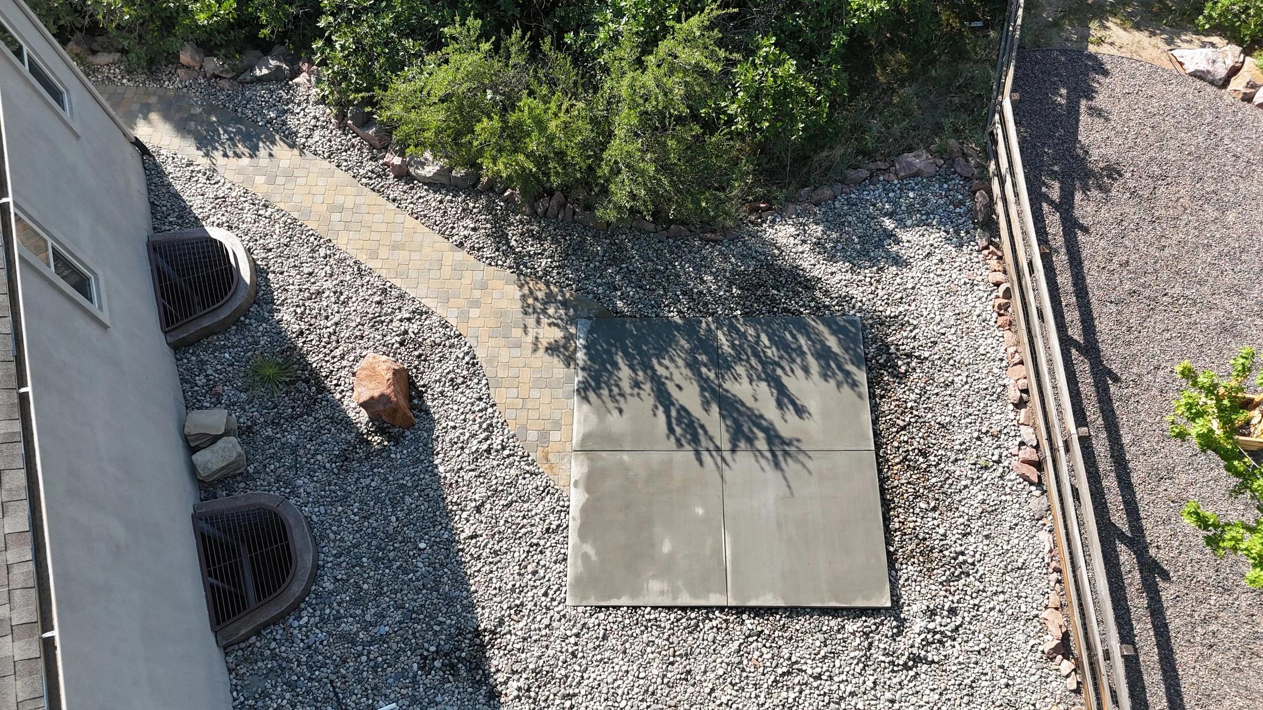 An aerial view of a backyard with a paved pathway, decorative rocks, small bushes, and green trees. Part of a concrete patio and a white house wall are visible on the left. A metal railing is on the right side of the yard.