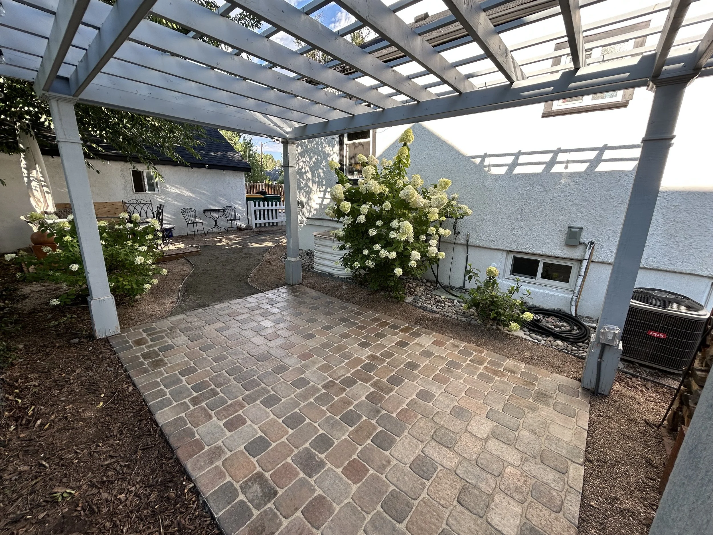 Backyard patio with brick flooring, a white garden shed, flowering shrubs, metal patio furniture, and a lattice pergola.