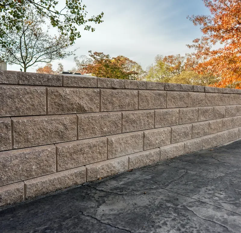 A beige brick wall with trees and autumn foliage in the background, and a concrete sidewalk in the foreground.