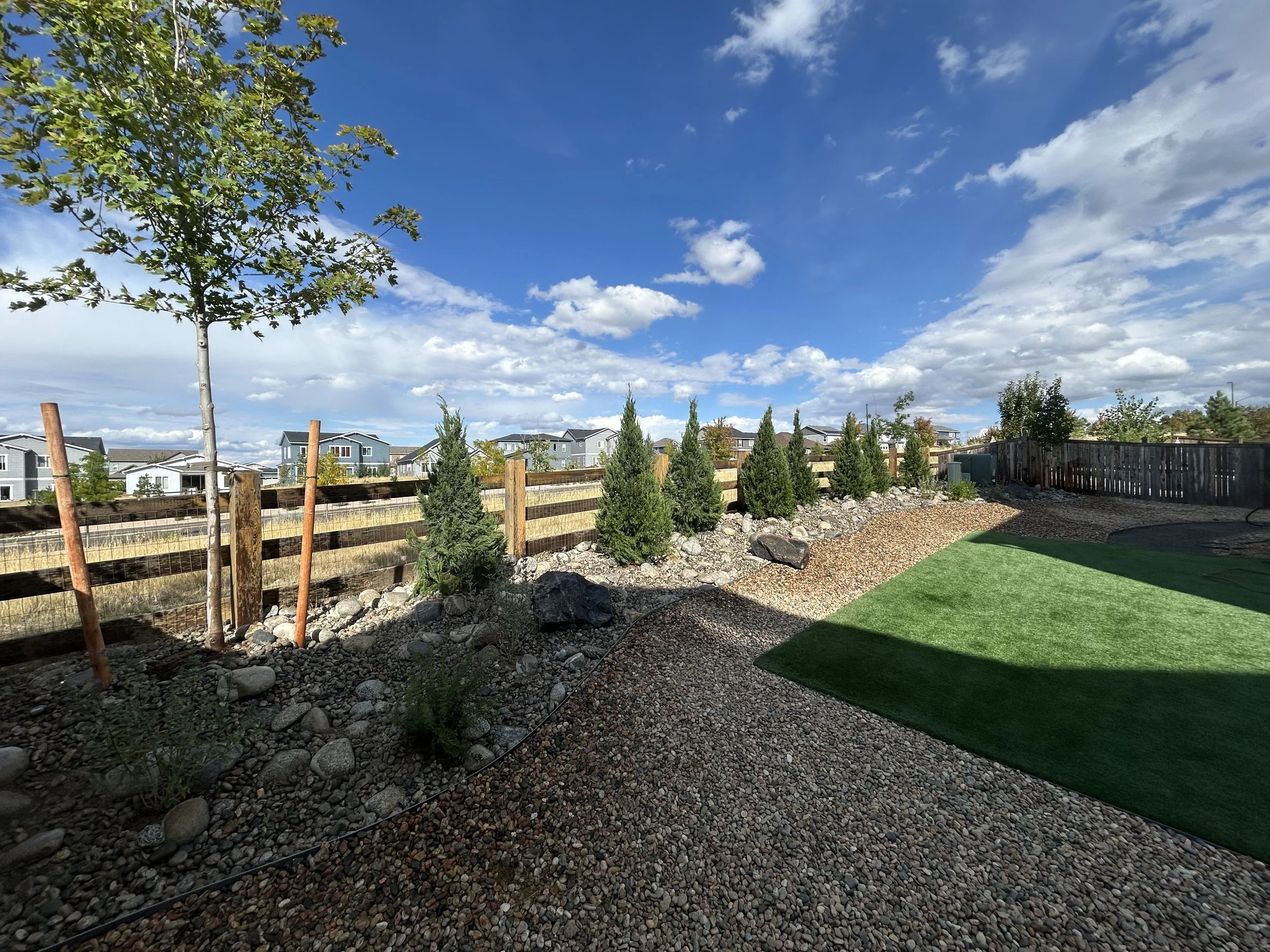 A backyard with a small green artificial lawn area, gravel pathway, young trees, and a wooden fence under a partly cloudy sky.