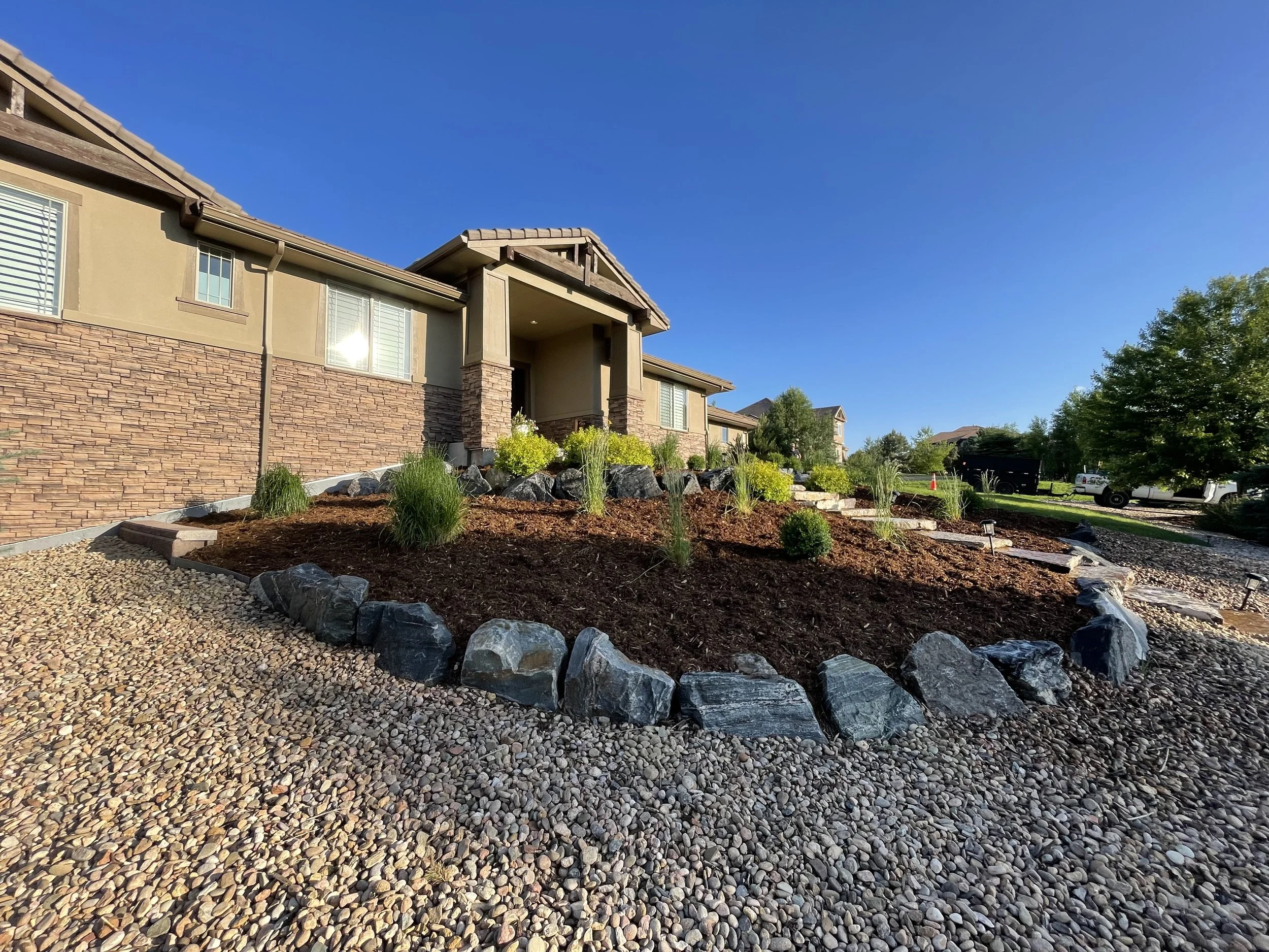 The front yard of a house with a landscaped garden, featuring rocks, mulch, small shrubs, and plants, with a clear blue sky and other houses in the background.