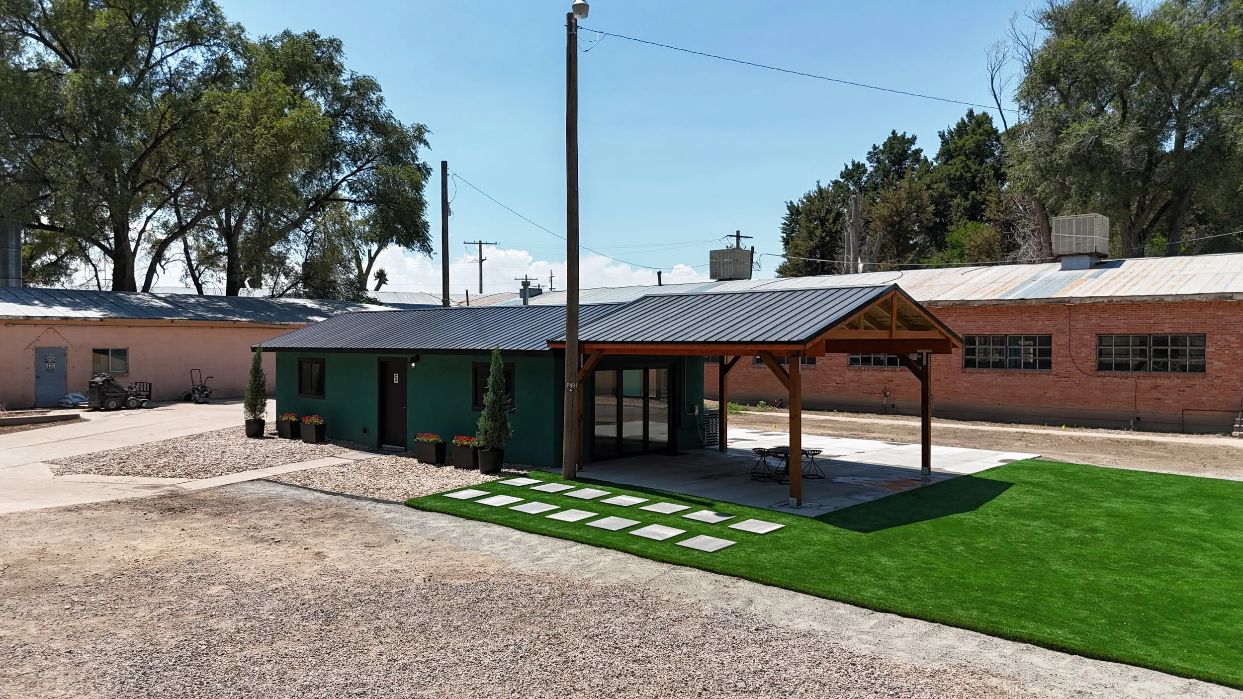 Modern small building with green exterior and metal roof, surrounded by artificial grass and concrete pavers, with outdoor seating, potted plants, and a covered porch, in a partly industrial area.